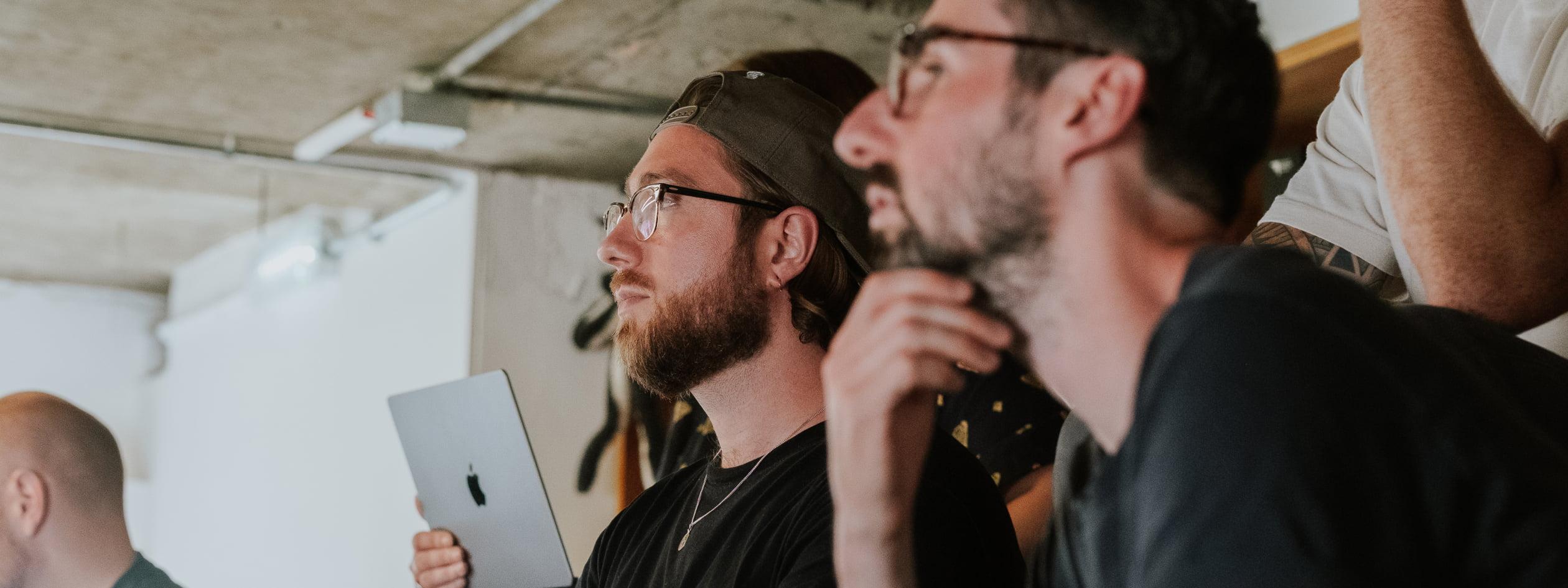 Two men look towards a presentation in a workshop setting, one holding a laptop, in a room with exposed concrete ceilings.