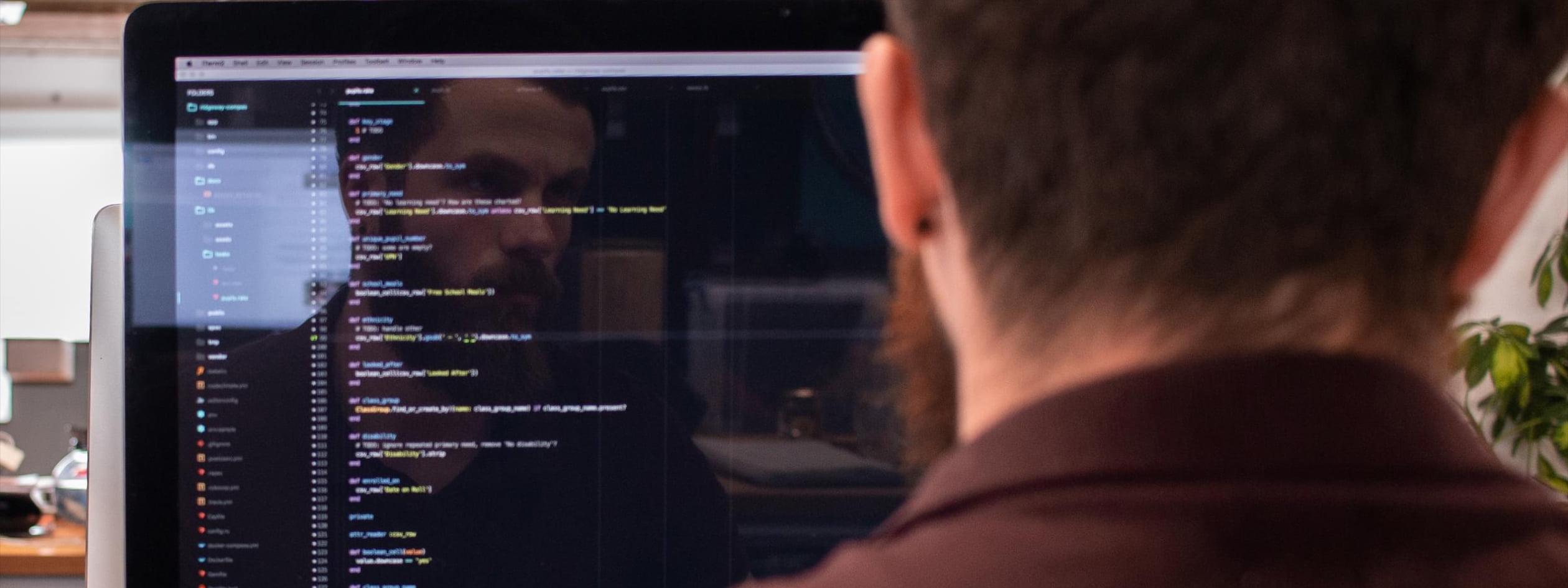 A close-up over the shoulder shot of a man working at a computer. On his screen is a code editor application.