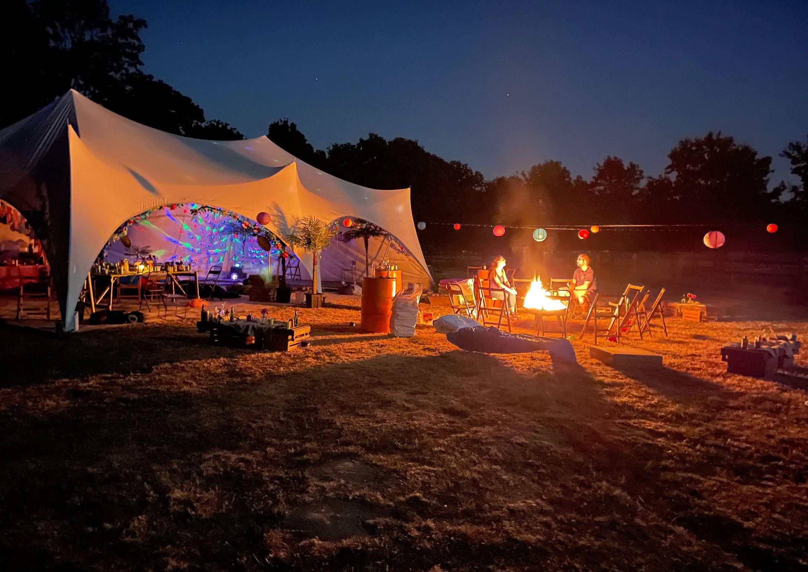 A nighttime outdoor gathering with a lit fire pit, festival-style tent, string lights, and people sitting around the fire in a field.