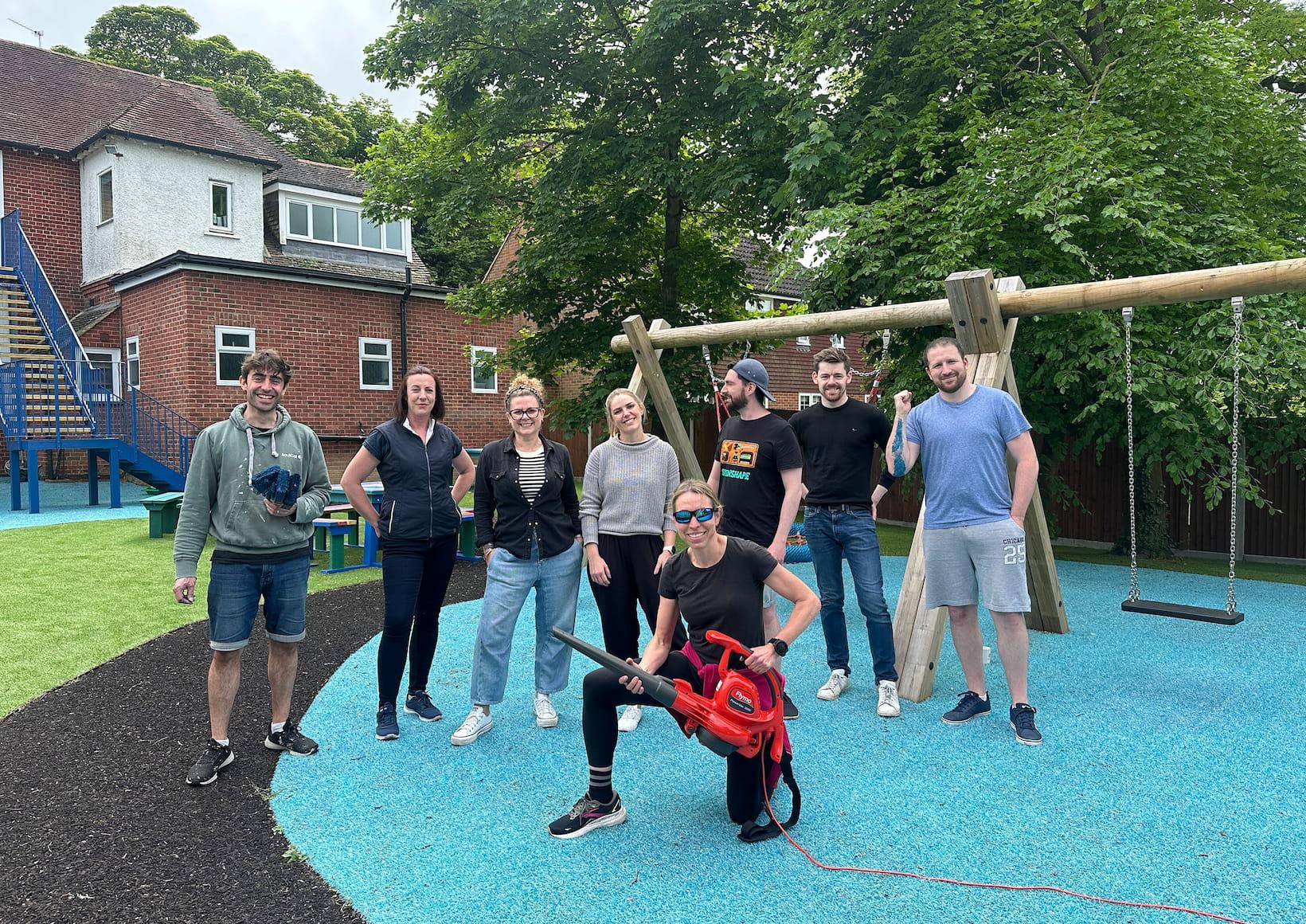 A group of volunteers posing in a playground they have been working on, with one person holding a leaf blower and others smiling behind them.