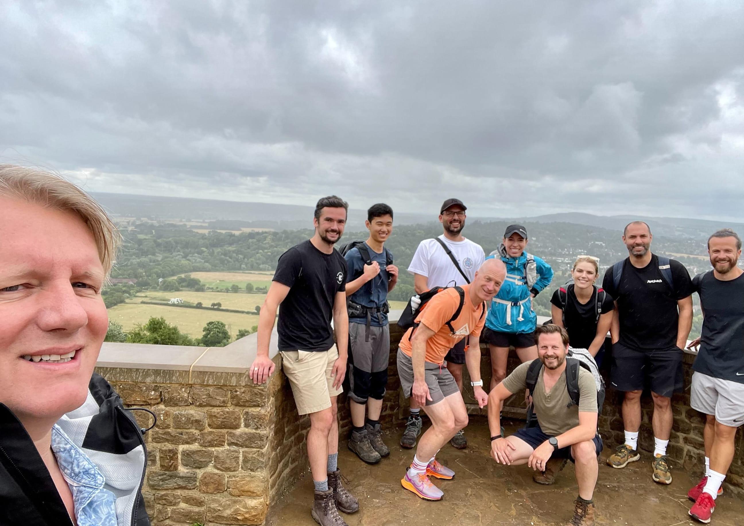 A group of hikers pose for a photo at a scenic hilltop viewpoint on a cloudy day, smiling and dressed in mixed hiking gear.