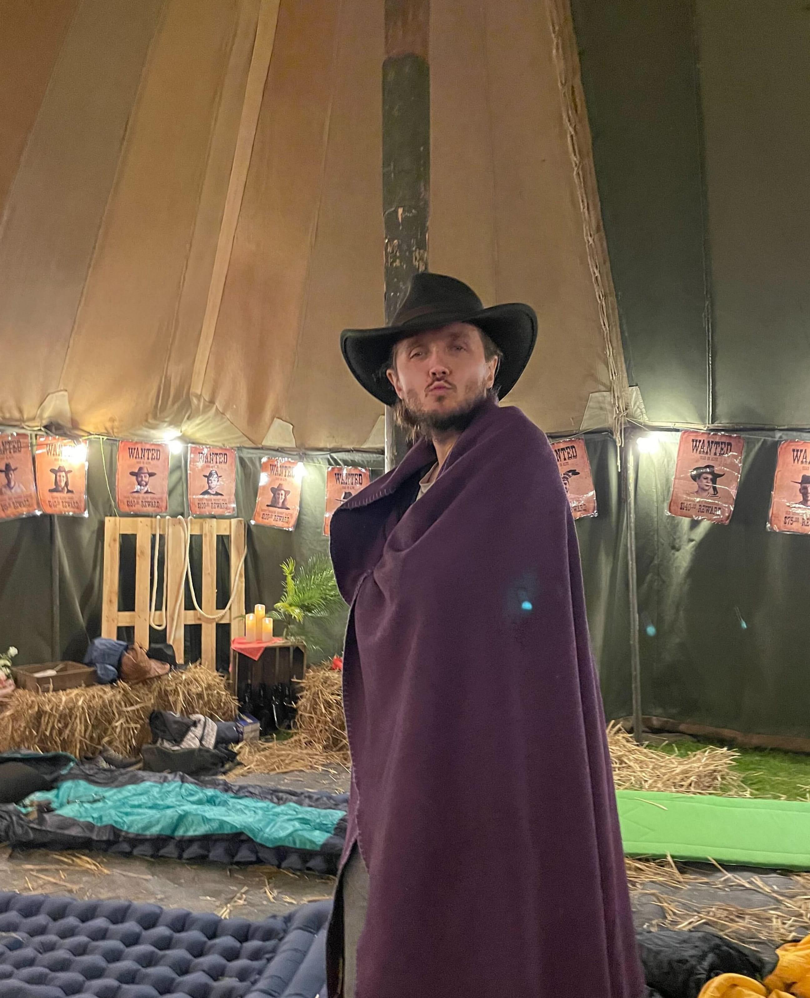 A person dressed in a cowboy hat and a purple blanket poses inside a tipi-style tent decorated with hay bales, “Wanted” posters, and warm string lights.
