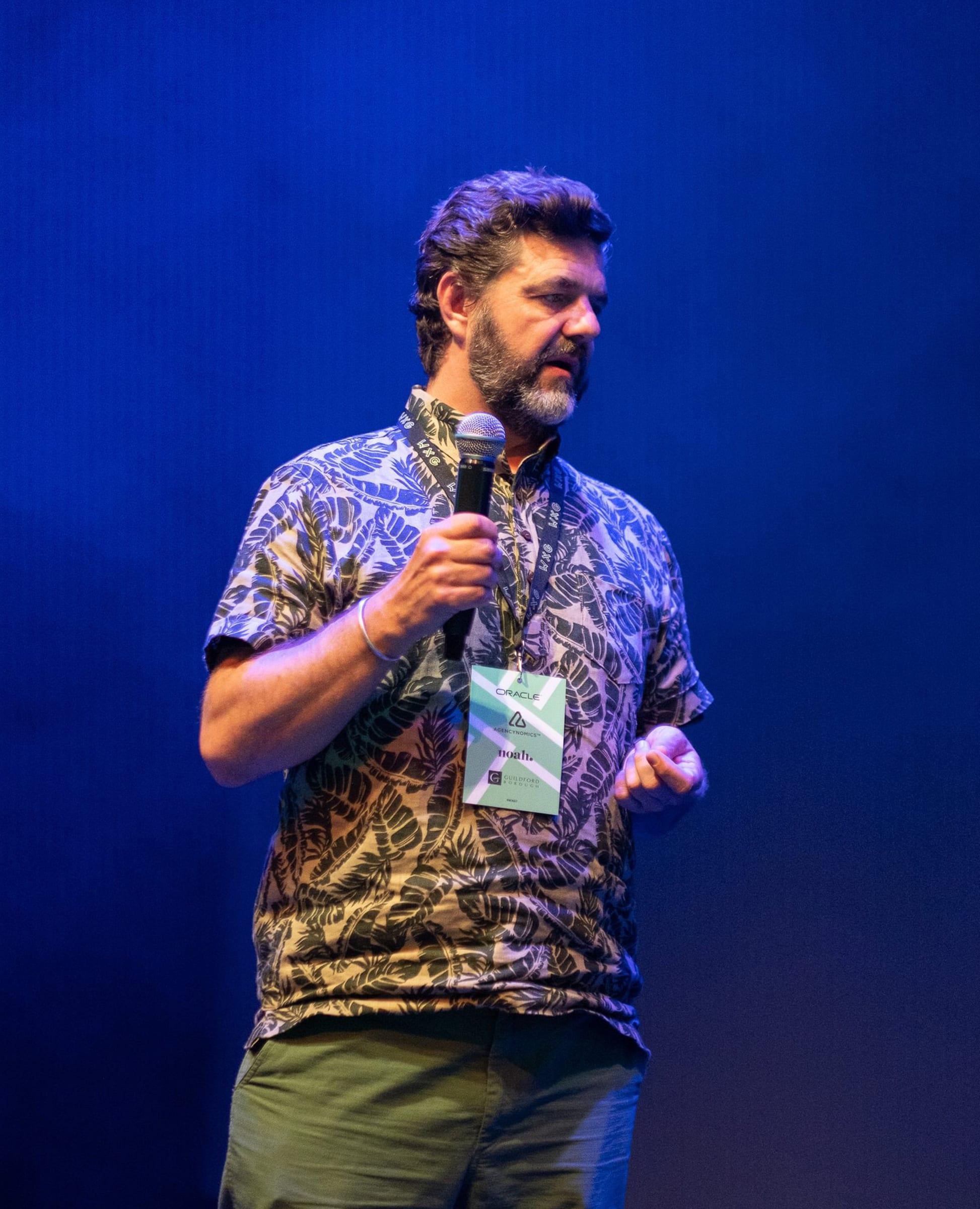 A man holding a microphone speaks on stage under blue lighting, wearing a patterned shirt and event lanyard.
