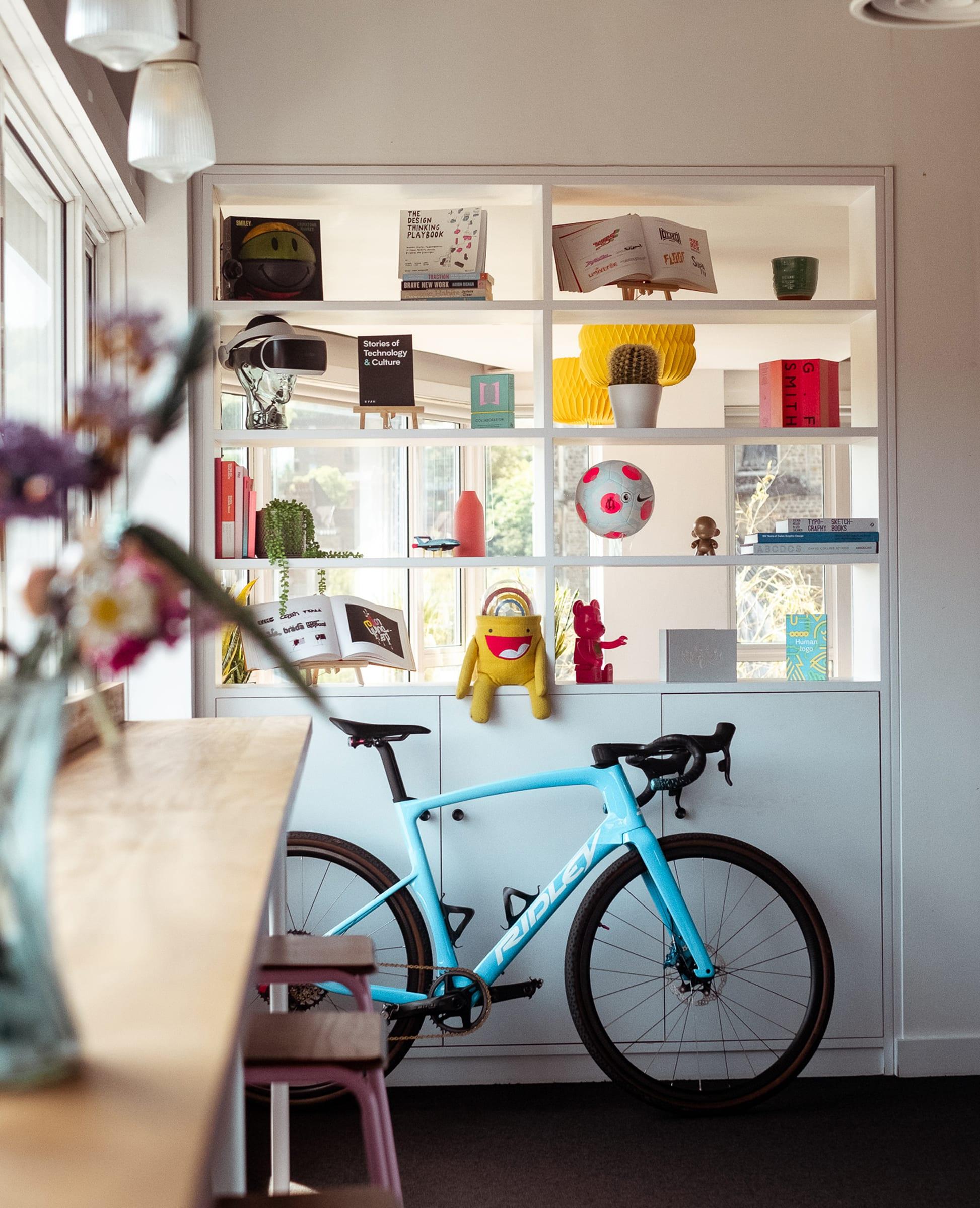 A turquoise road bike parked inside an office, next to a shelving unit filled with books and decorative items.