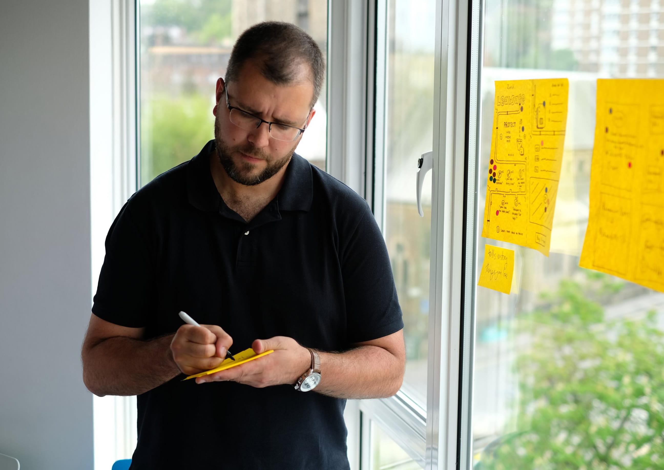A man writes on a yellow sticky note while standing by a window, with other sketches and notes taped to the glass.