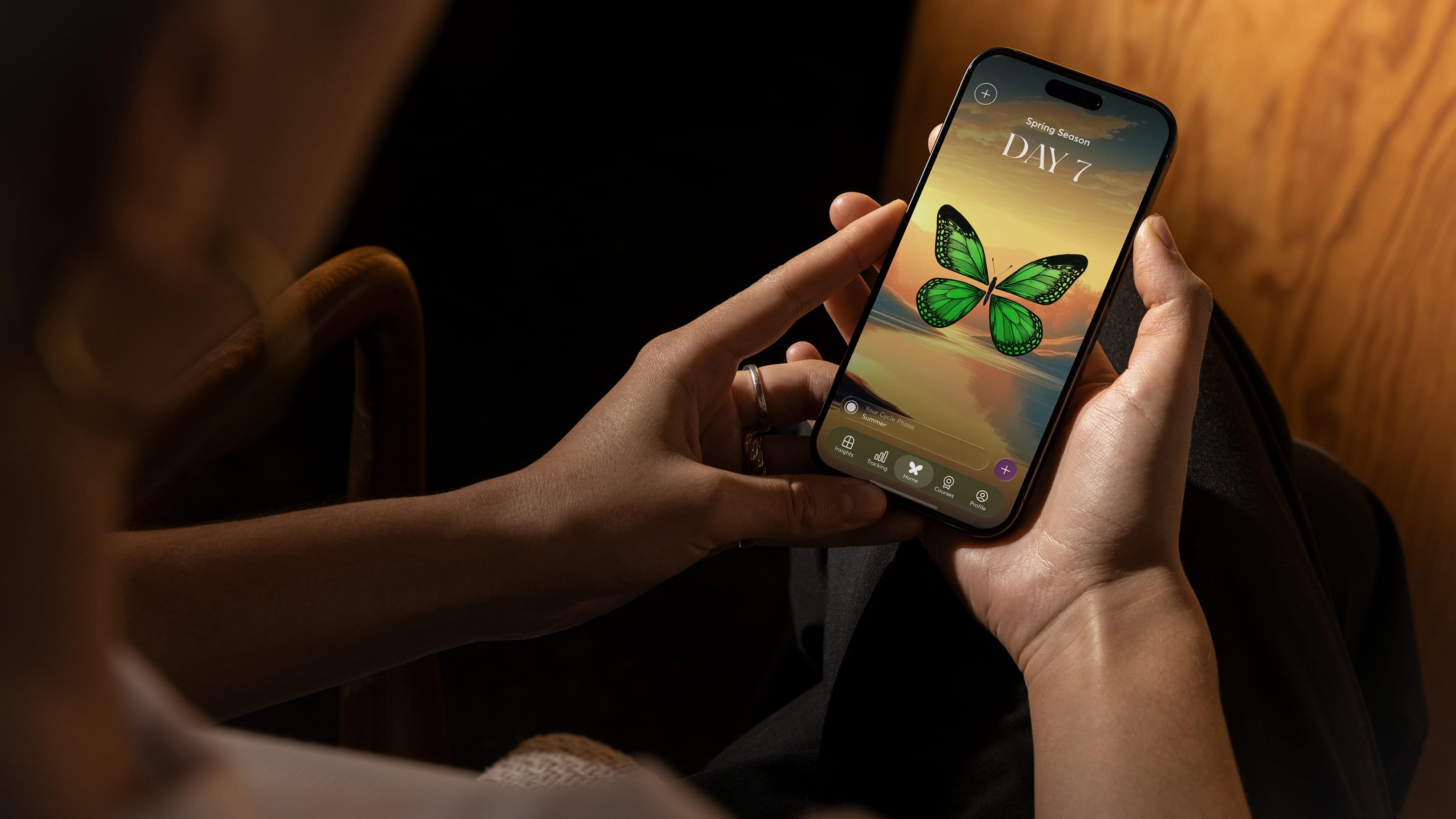 Hands holding an iPhone showing the 28X app, with a green butterfly companion over a sunset landscape on the 'Spring Season, Day 7' home screen.
