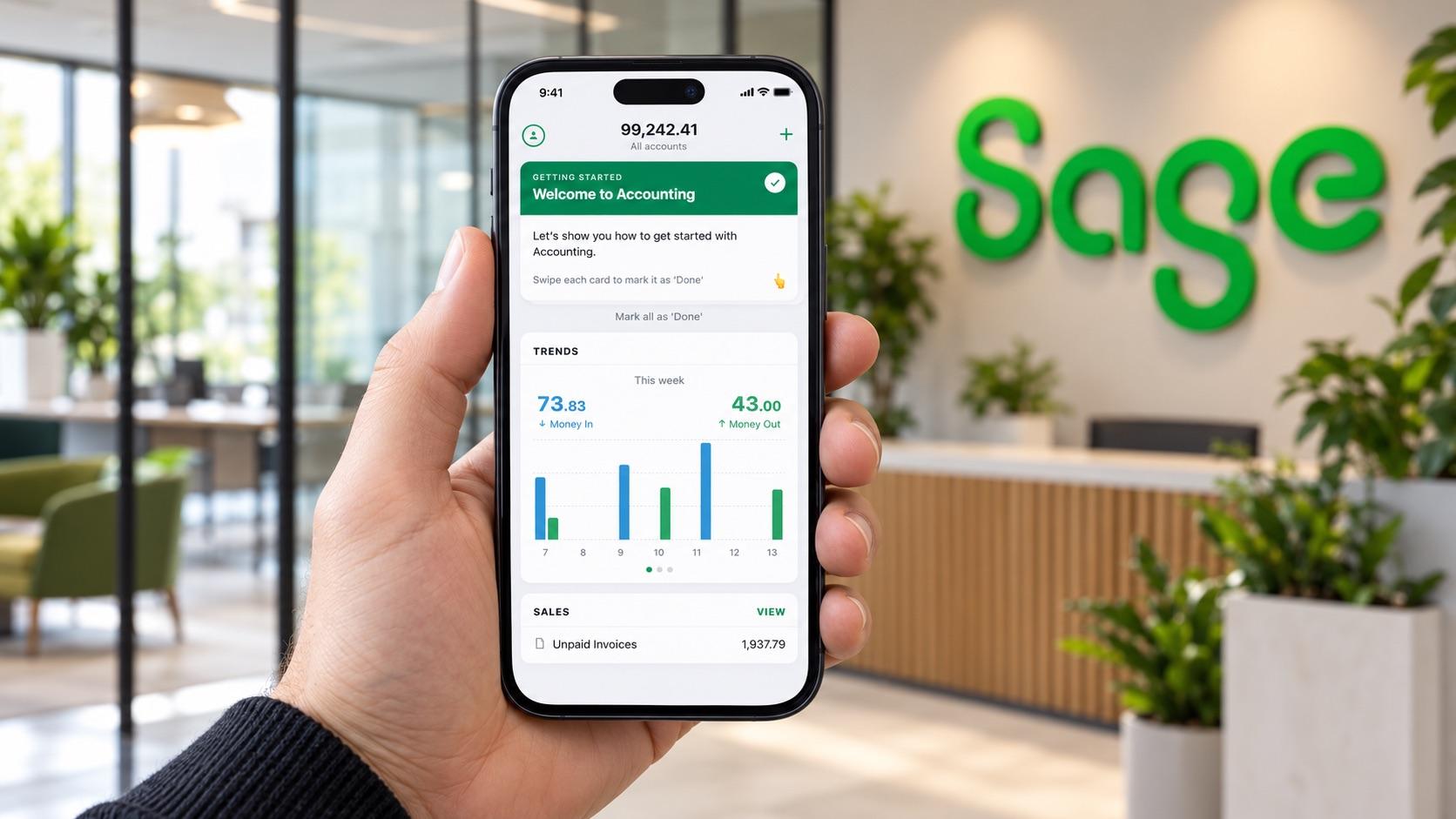 A hand holding a smartphone showing the Sage Accounting app's welcome screen and weekly money in and out chart, with the green Sage logo on the office wall behind.