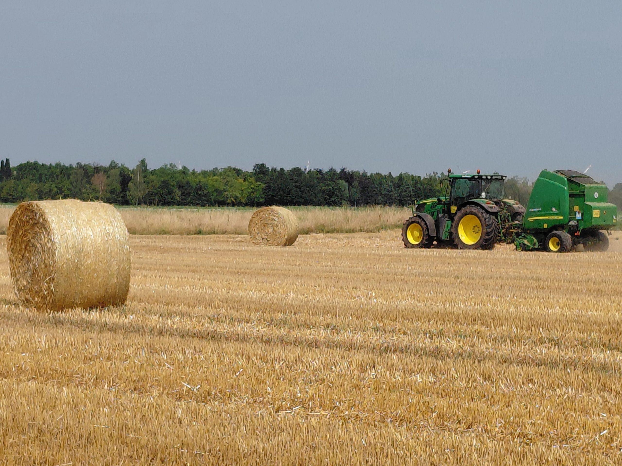 A green tractor and baler working a harvested field dotted with large round straw bales under a hazy sky.