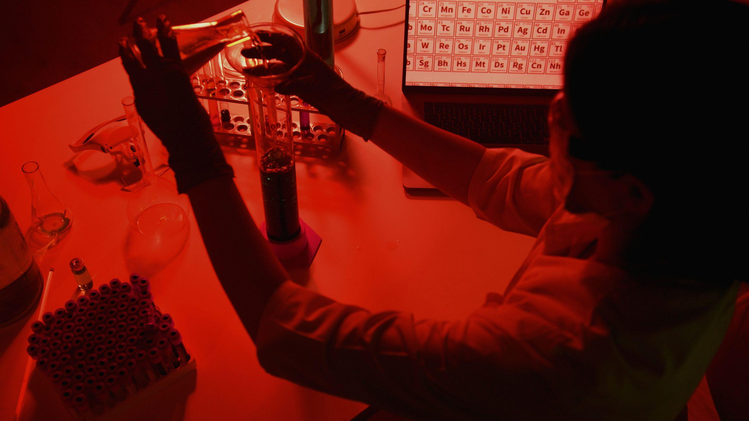 A scientist in gloves pouring a sample into a test tube under red laboratory light, with a periodic table on a screen behind.