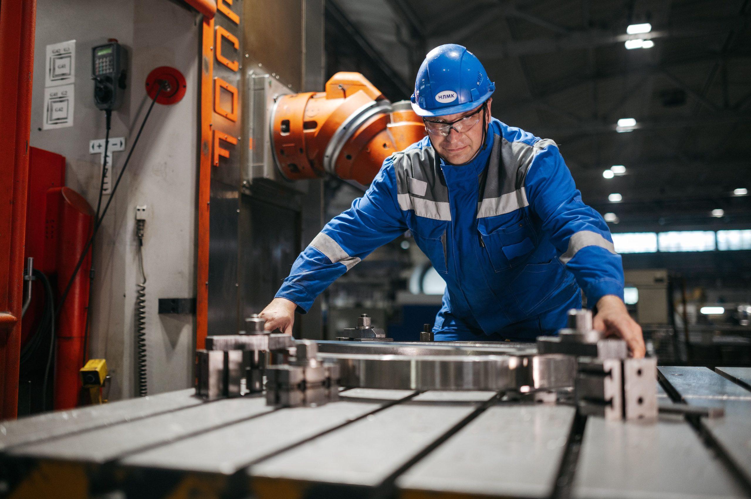 A worker in a hard hat and safety glasses operating machinery to position a large steel component in a workshop.
