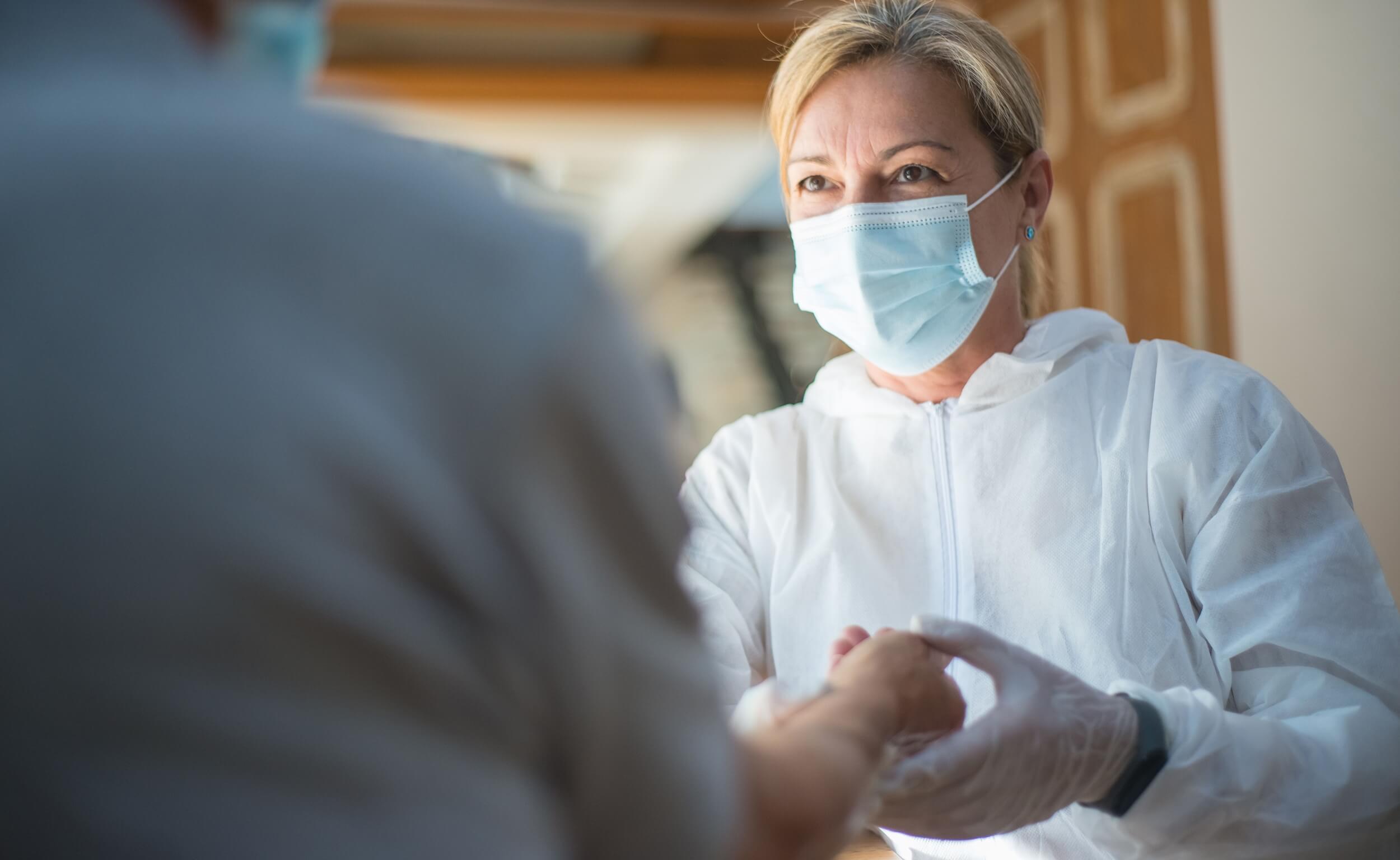 A healthcare worker in a mask and protective clothing holds the hand of an elderly patient.
