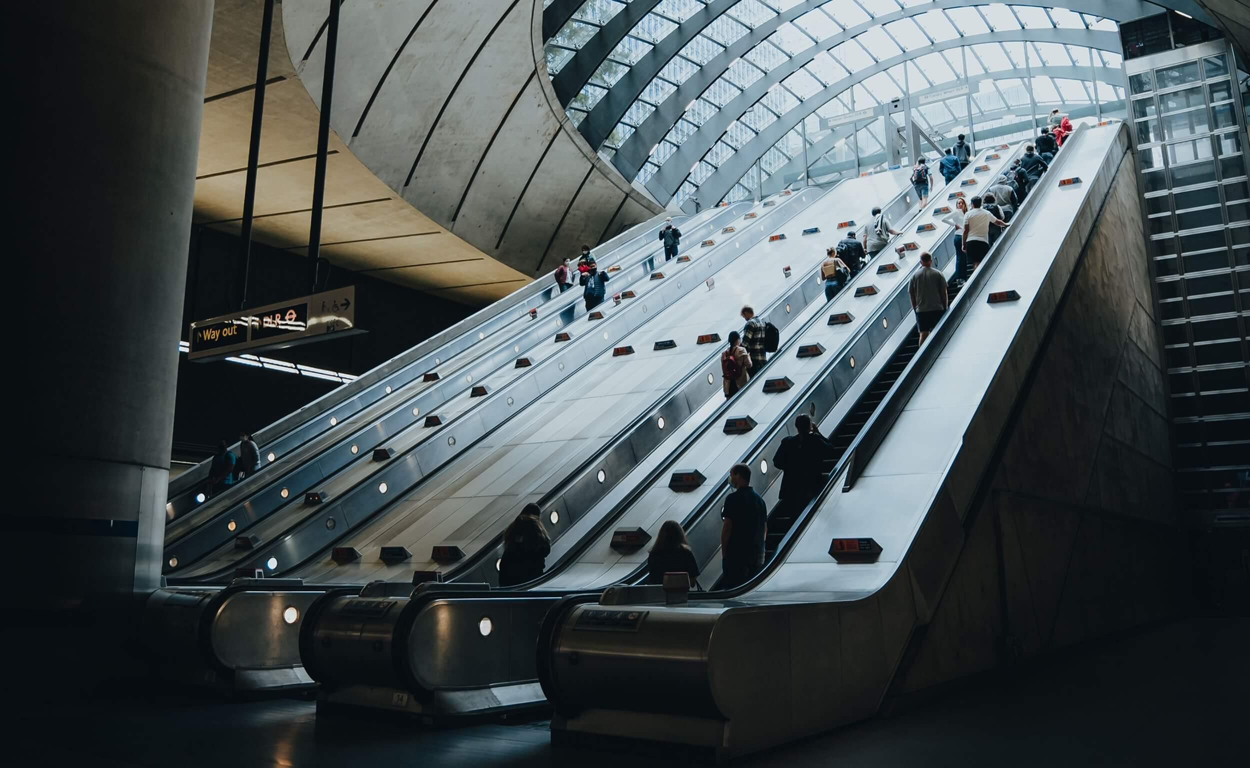 Commuters riding a wide bank of escalators beneath the glass-domed canopy of a modern underground station.