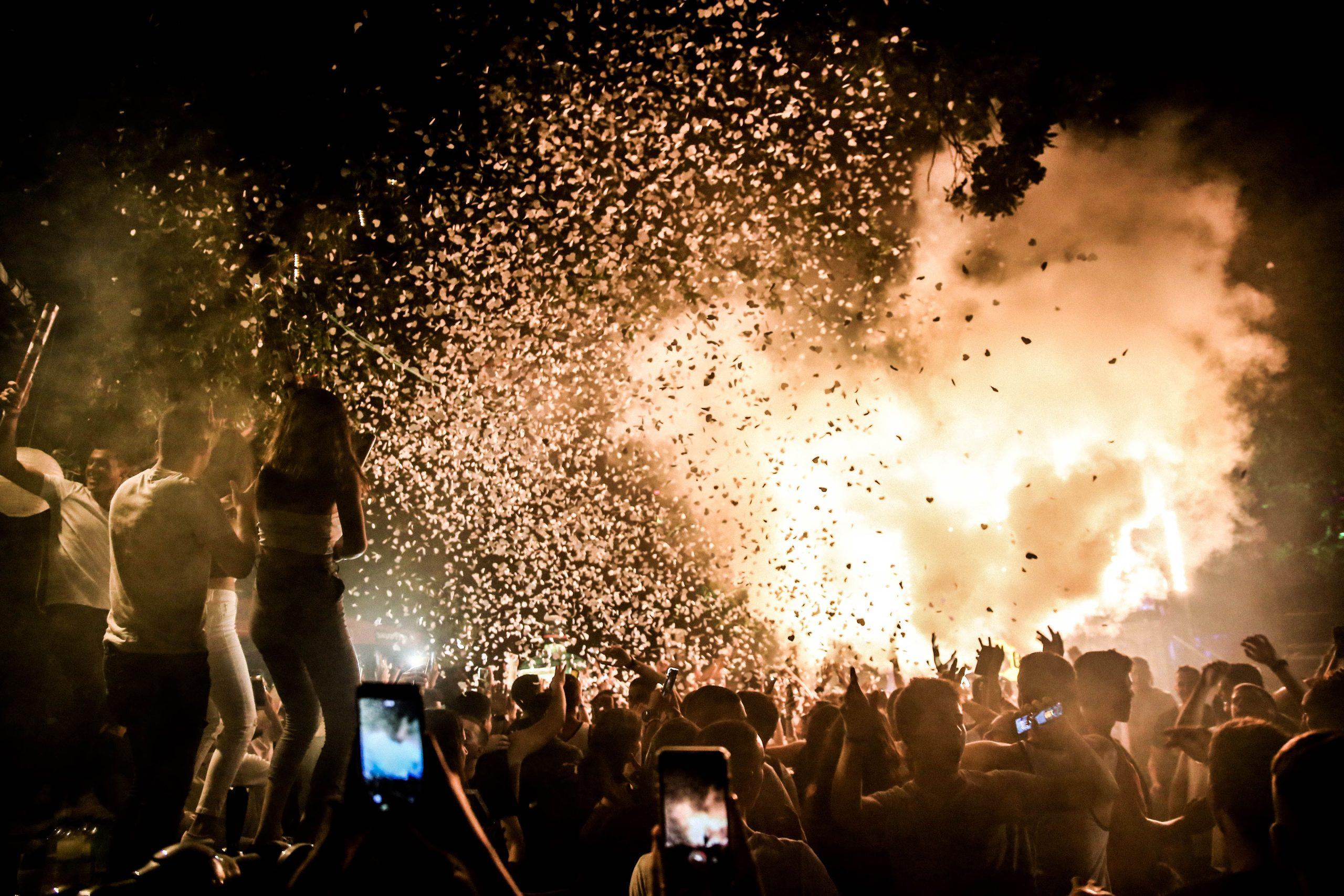 A festival crowd at night filming a confetti-filled stage moment on their phones.