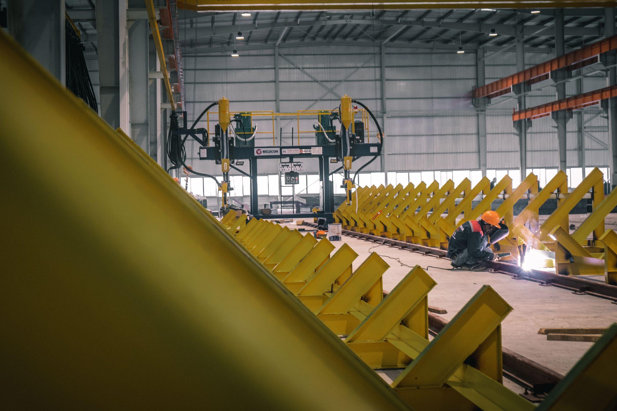A worker welding steel components among rows of bright yellow fabricated metal in an industrial workshop.