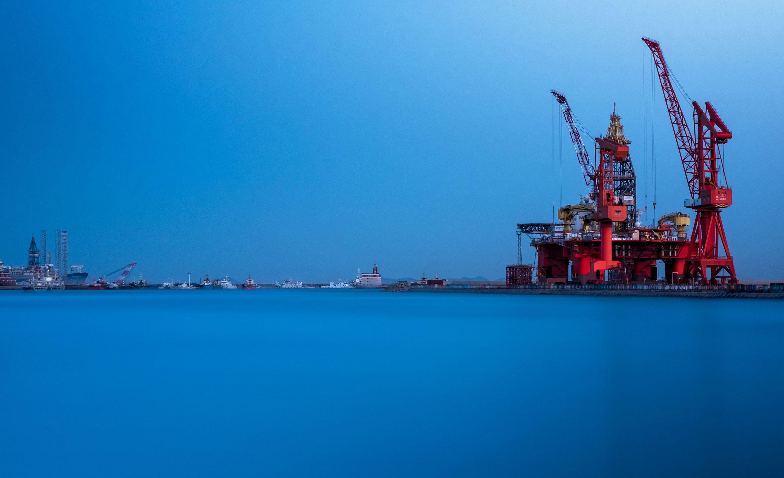 An offshore oil rig and cranes moored at a harbour under a deep blue dusk sky.