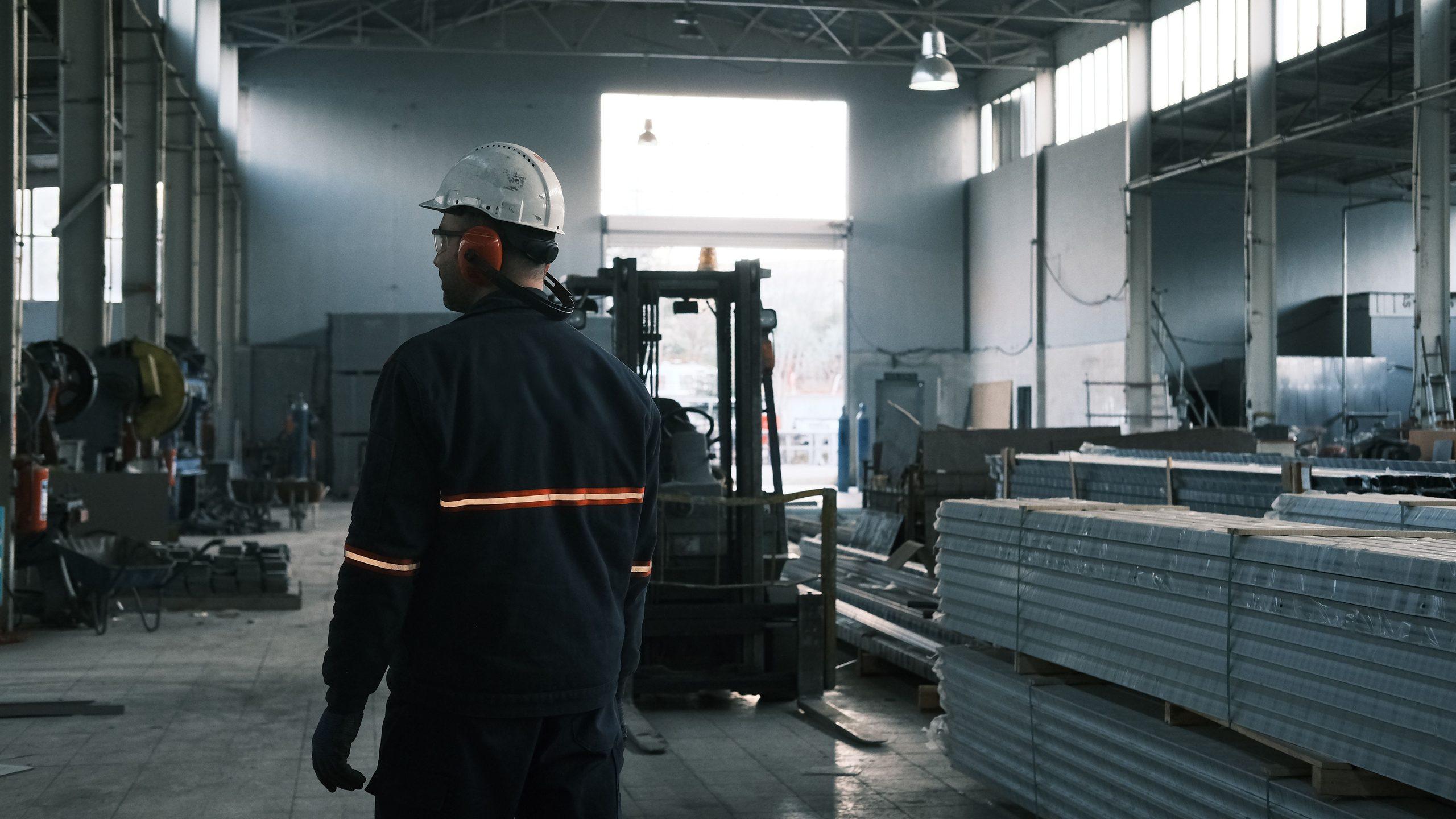A worker in a hard hat and hi-vis walking through a warehouse past stacked packaged materials and a forklift.
