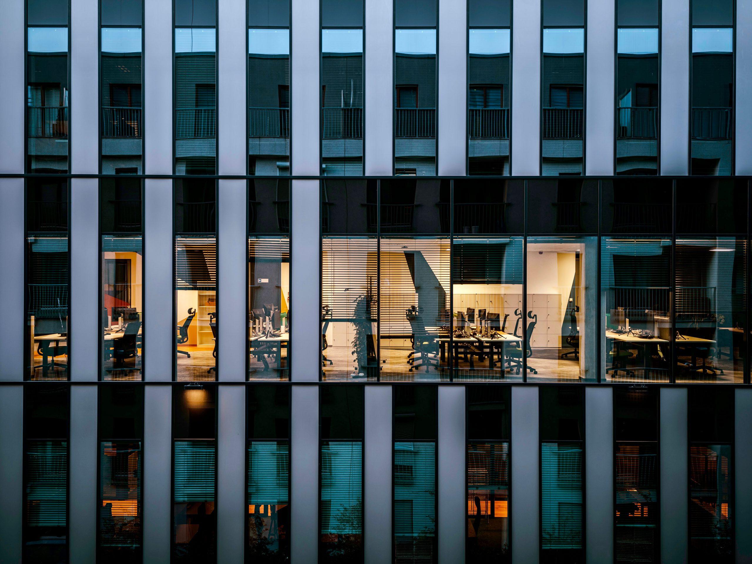 The glass facade of a modern office building at dusk, revealing lit open-plan workspaces inside.