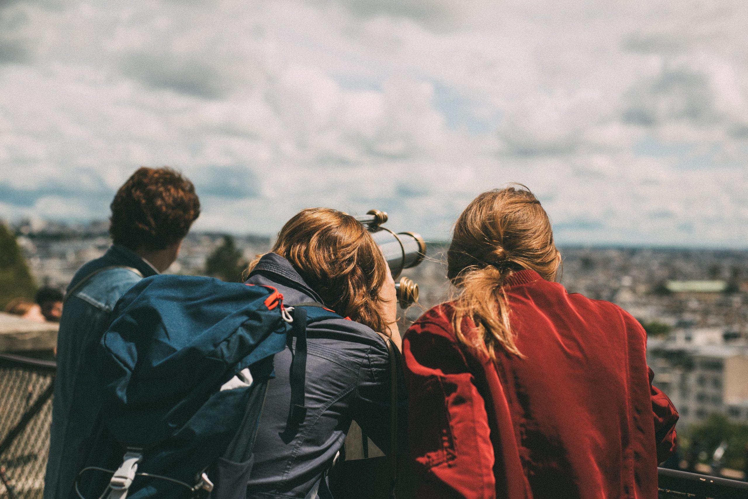 Three travellers with backpacks looking out over a city through a viewing telescope on a cloudy day.