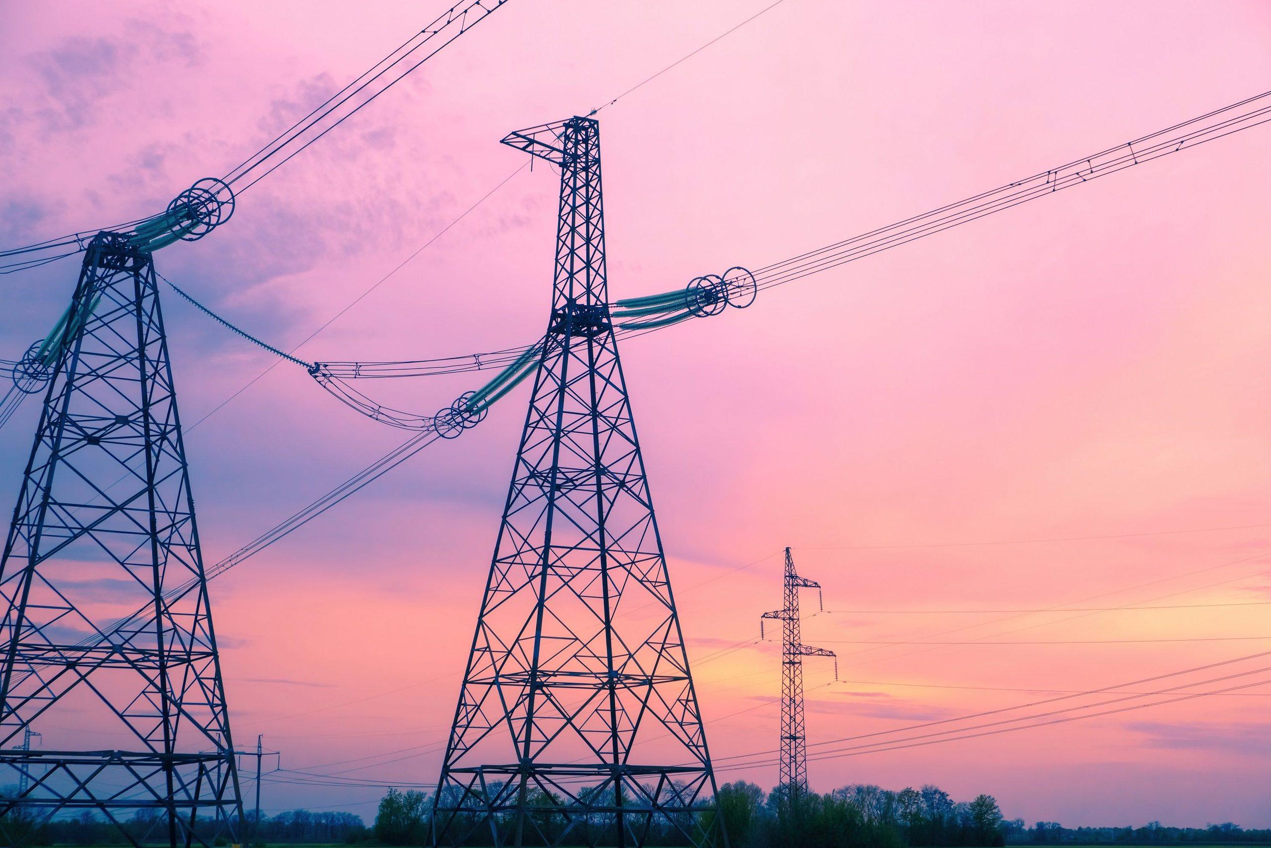 Electricity transmission pylons and power lines silhouetted against a pink and orange sunset sky.