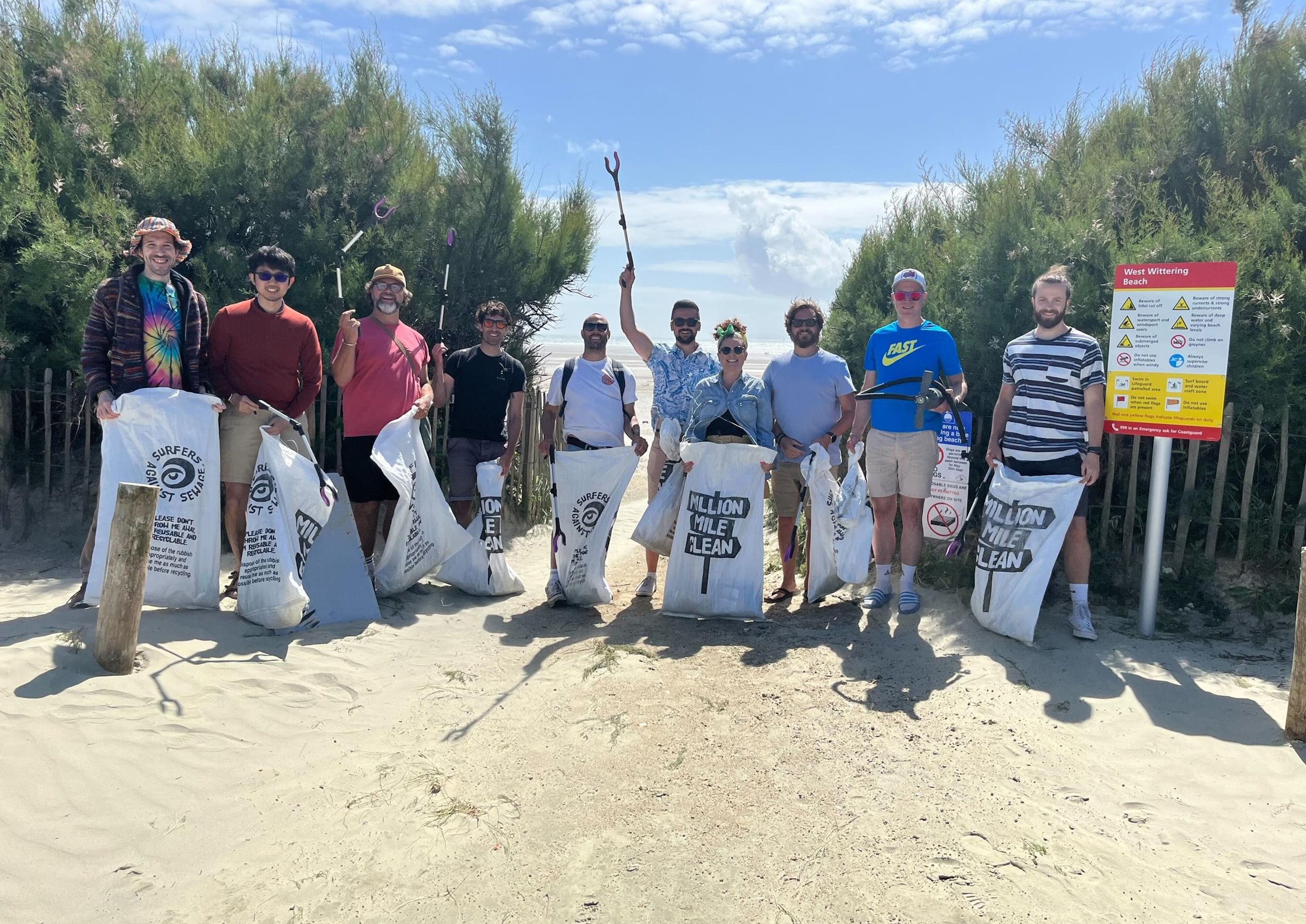 A group of people standing on a sandy path at West Wittering Beach, holding litter-picking bags and tools after a beach clean, smiling in front of green bushes under a bright sky.