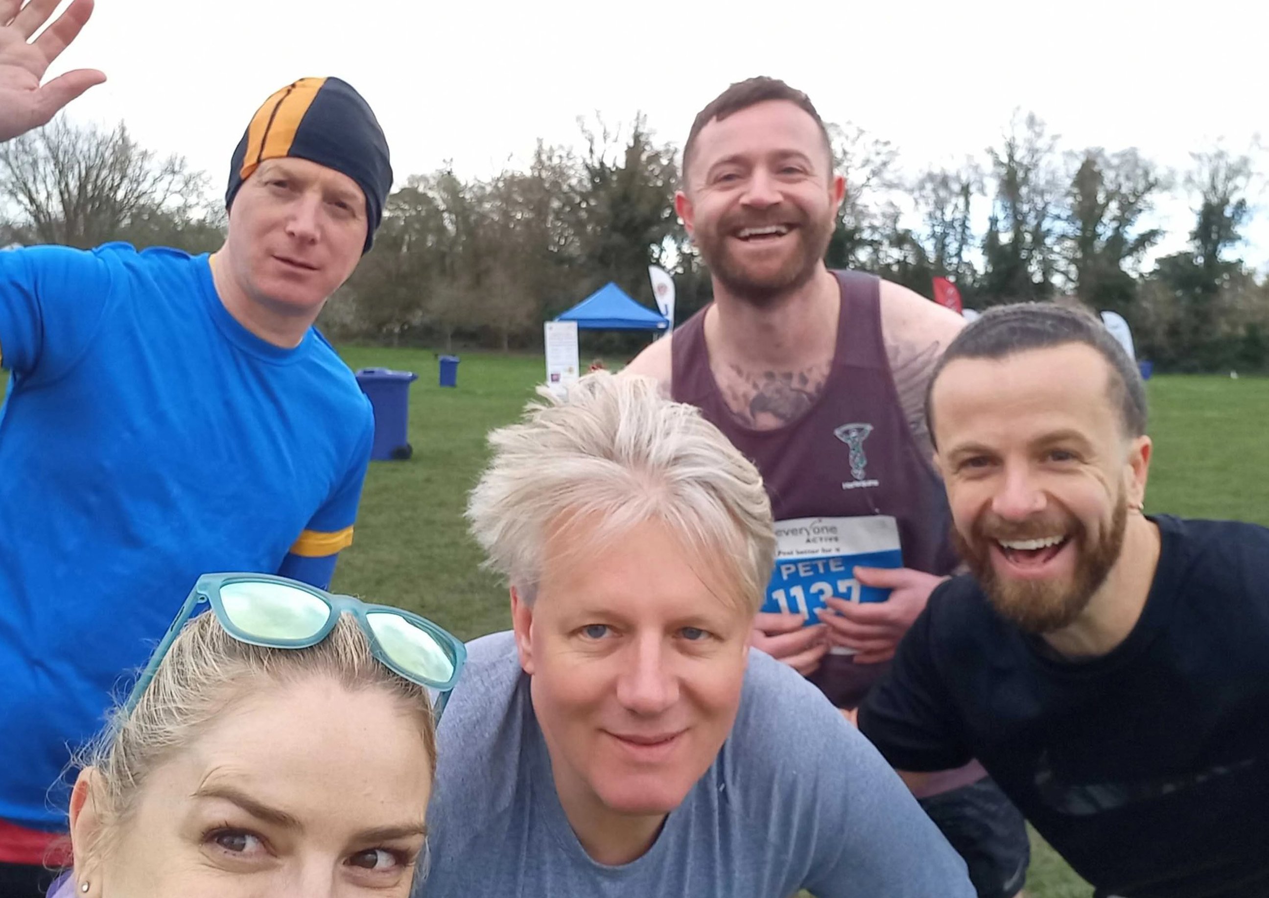 Five runners posing and smiling for a selfie at an outdoor running event, with trees and event tents in the background.