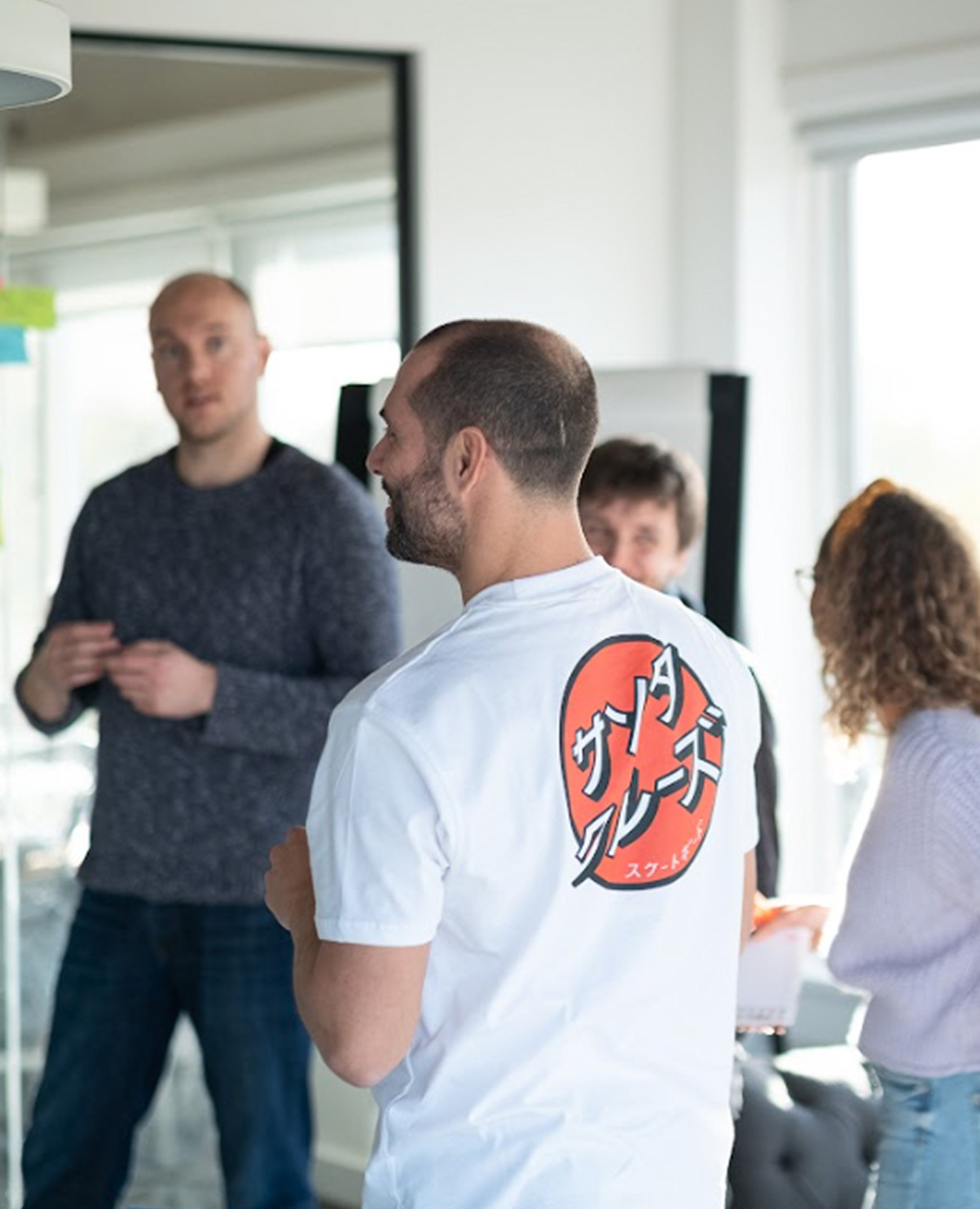 Team members chatting in a bright office space, with a man in a white graphic T-shirt facing away from the camera while others stand around discussing.