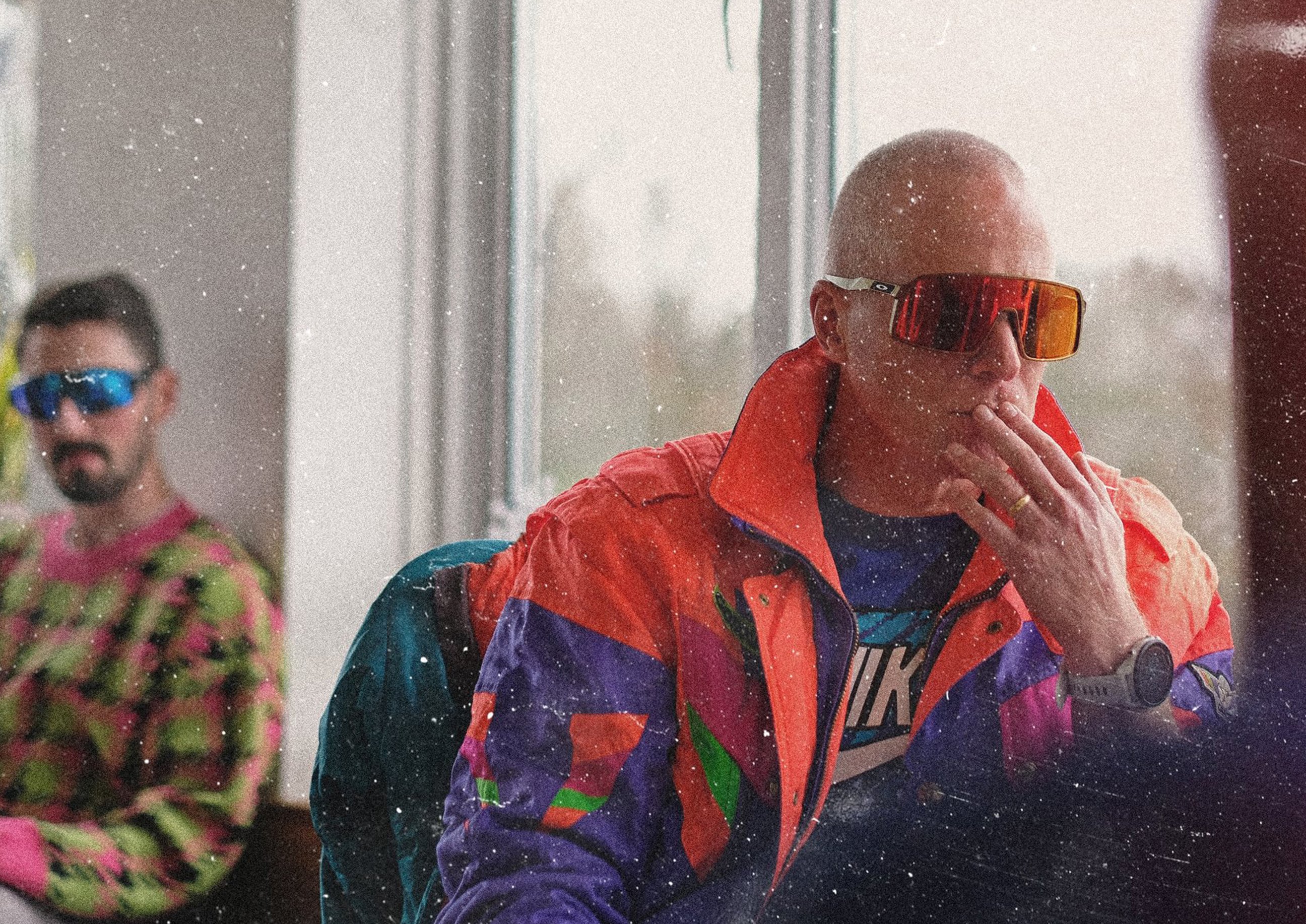 A man wearing oversized reflective sunglasses and a colourful retro windbreaker sits indoors with dramatic lighting, while another person in bright patterned clothes is blurred in the background.
