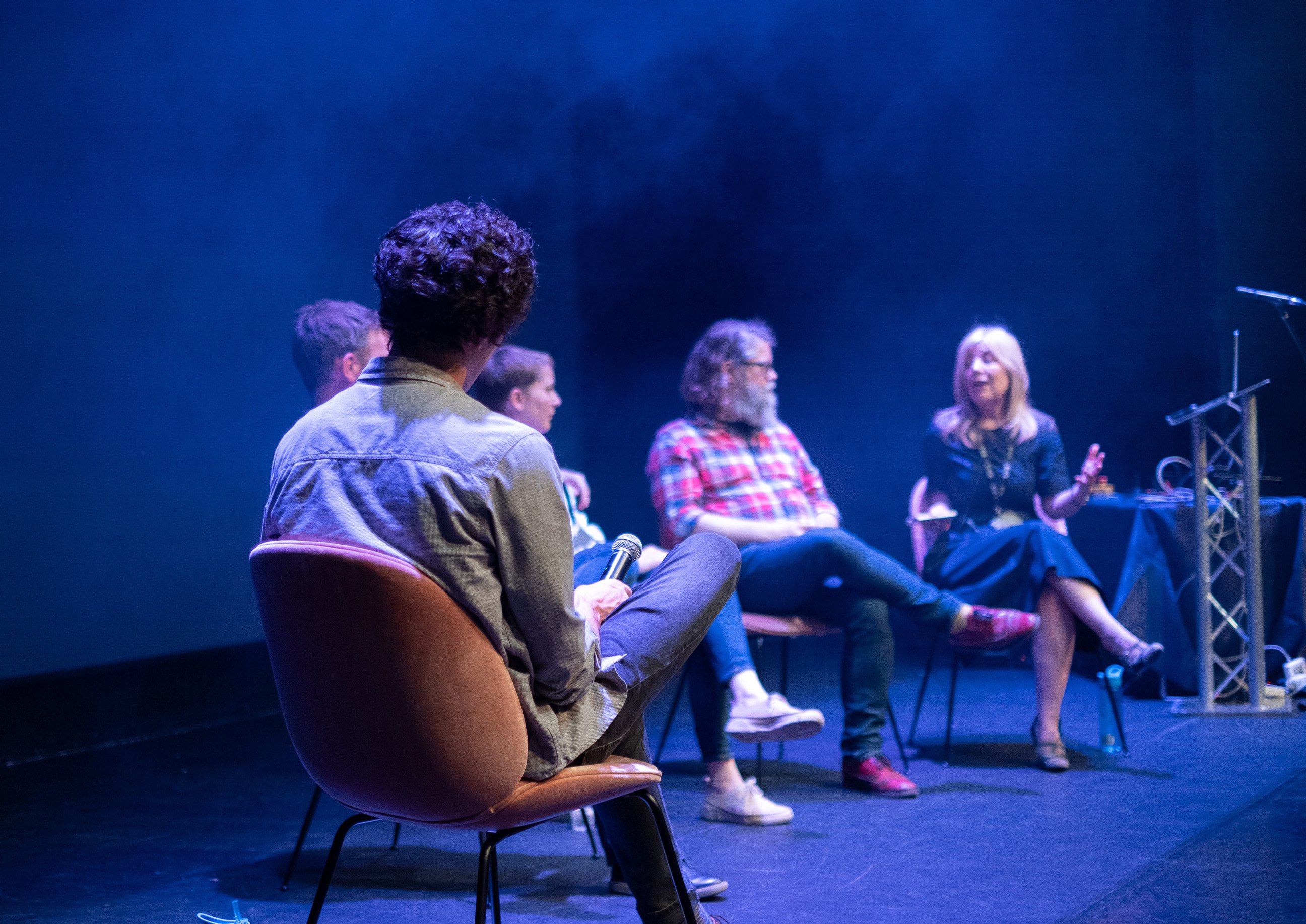 A panel of speakers seated on stage in soft blue lighting, engaged in discussion, with the audience’s view from behind one of the seated participants.