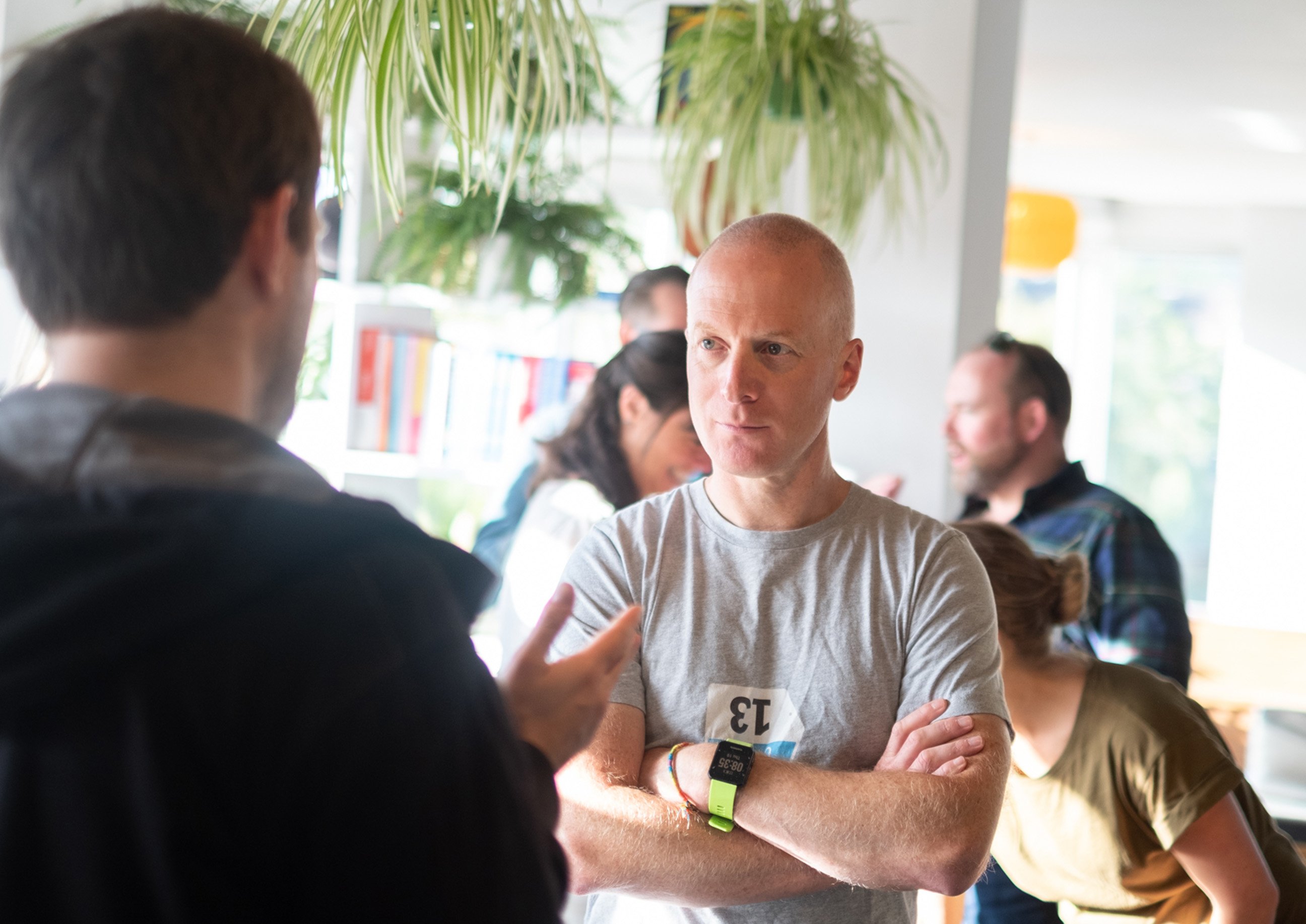 A man in a grey T-shirt listens intently with arms crossed during a conversation at a bright indoor gathering, with other people talking in the background and hanging plants overhead.