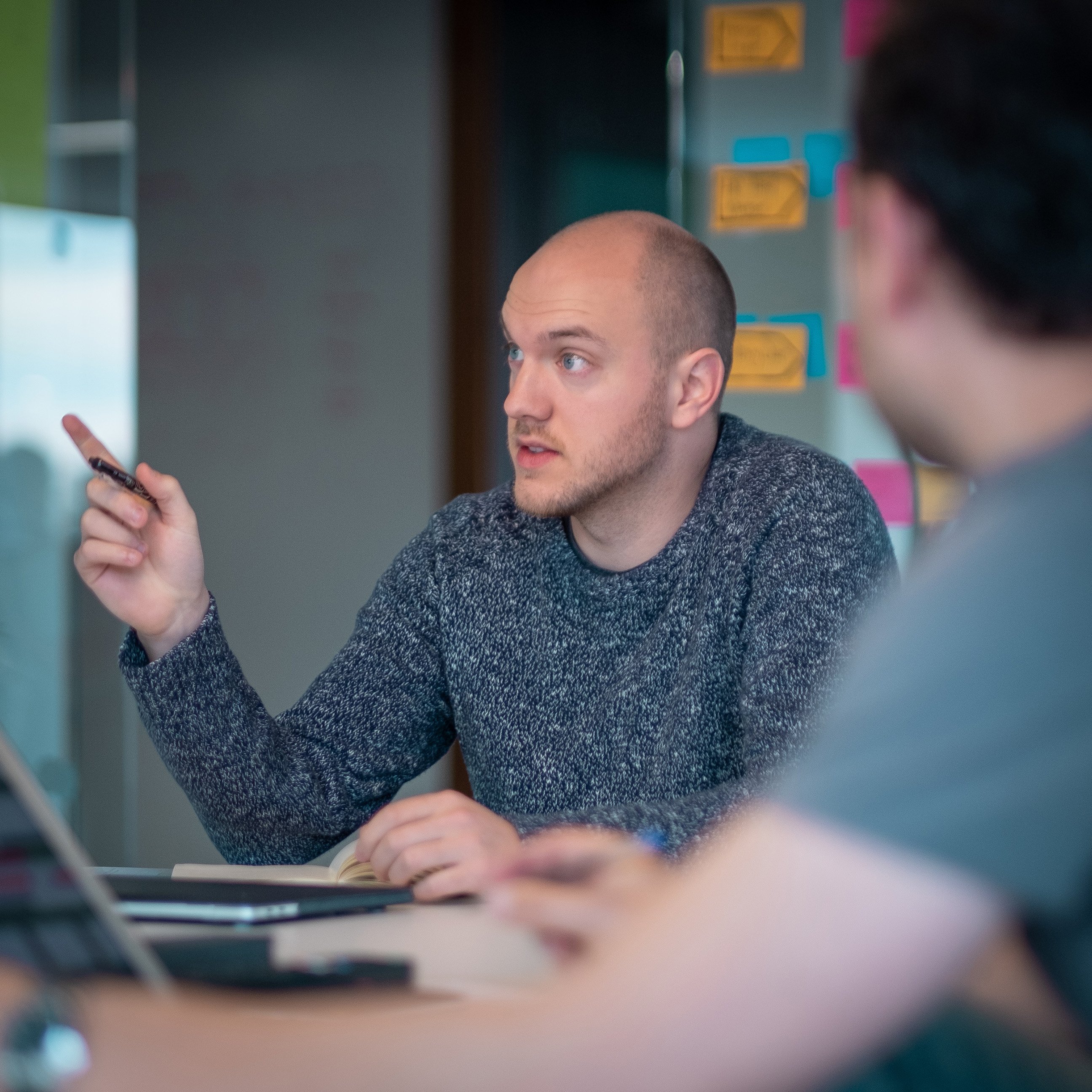 A man in a grey jumper speaks and gestures with a pen during a meeting, with colourful sticky notes on the wall behind him.”