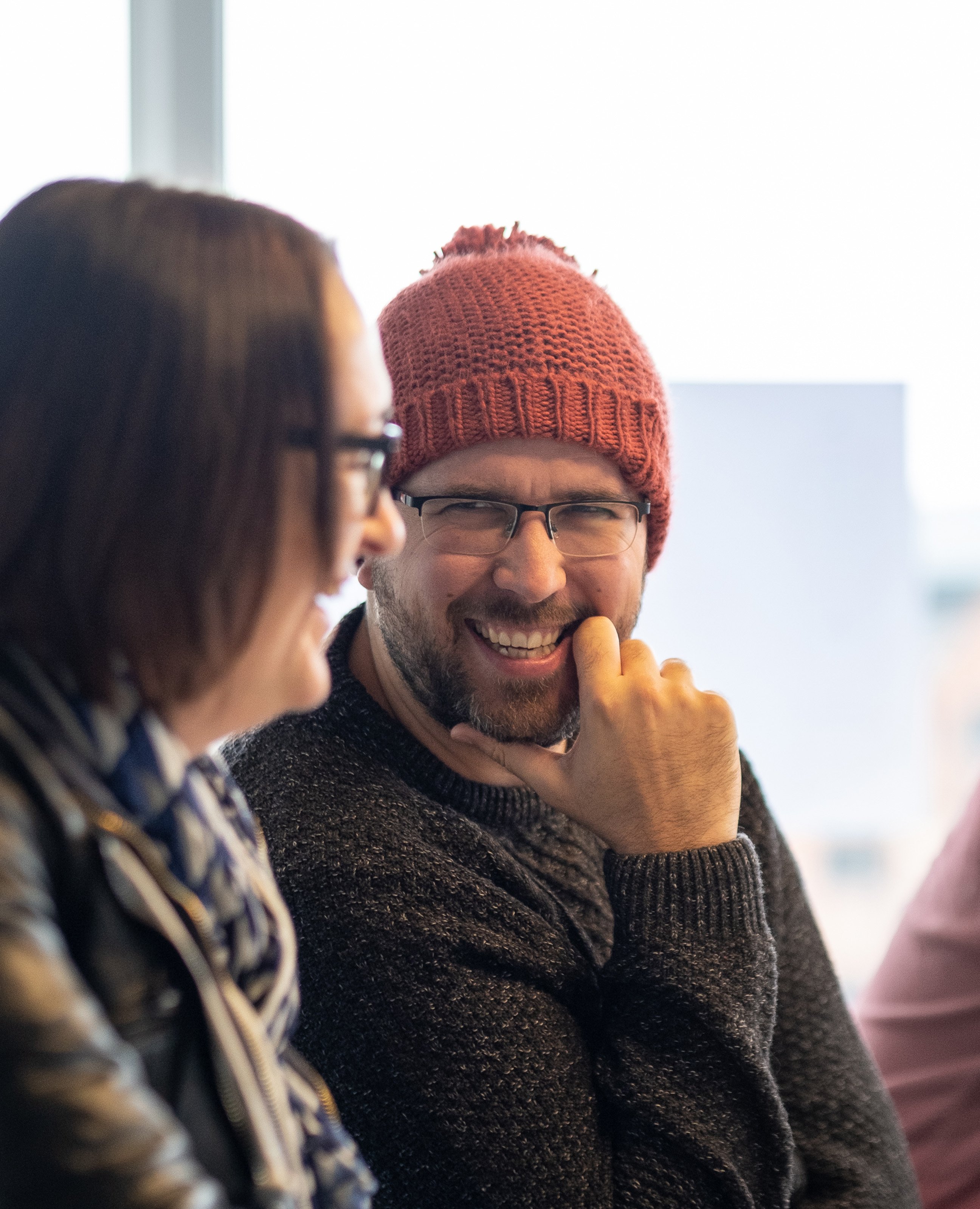 A man wearing a knitted red beanie smiles warmly while talking with a colleague during an indoor meeting.