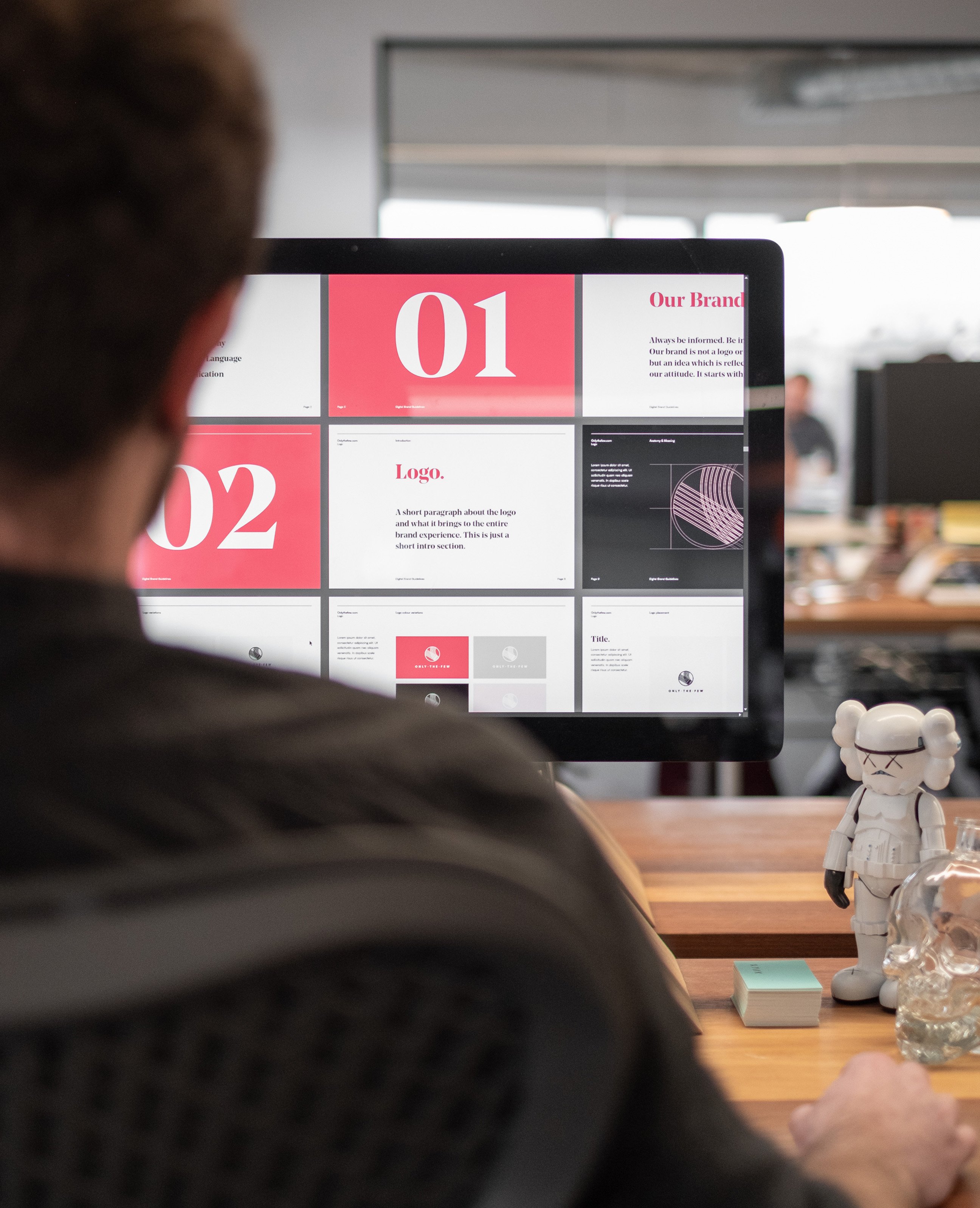 A designer sits at a desk reviewing brand guideline slides on a large monitor, with a robot figurine and glass skull ornament beside the screen.”