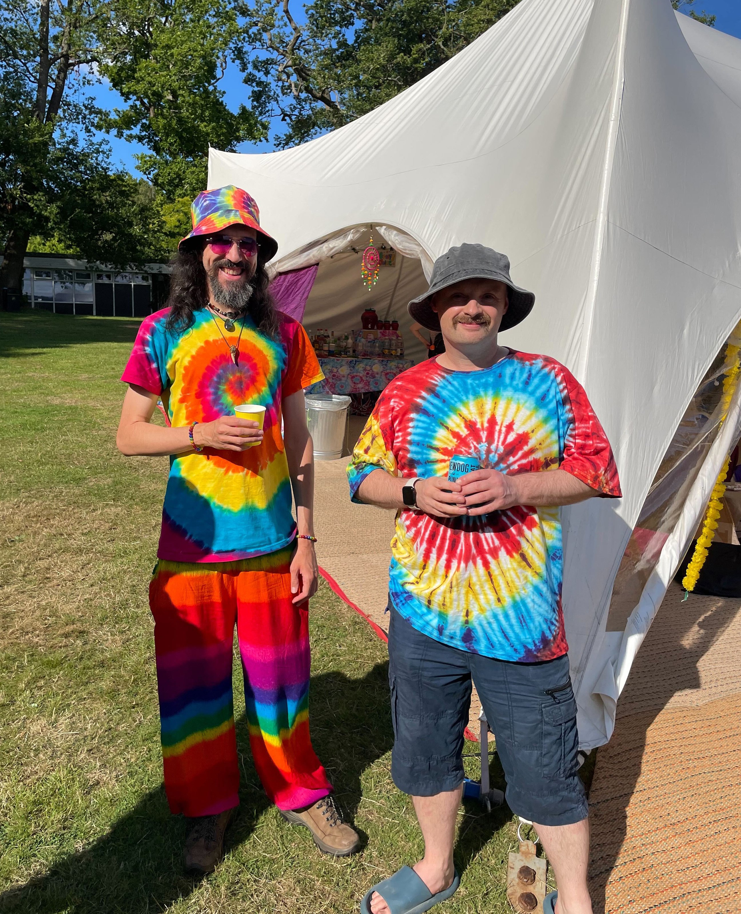 Two people wearing bright tie-dye outfits and bucket hats stand smiling outside a festival tent on a sunny day.