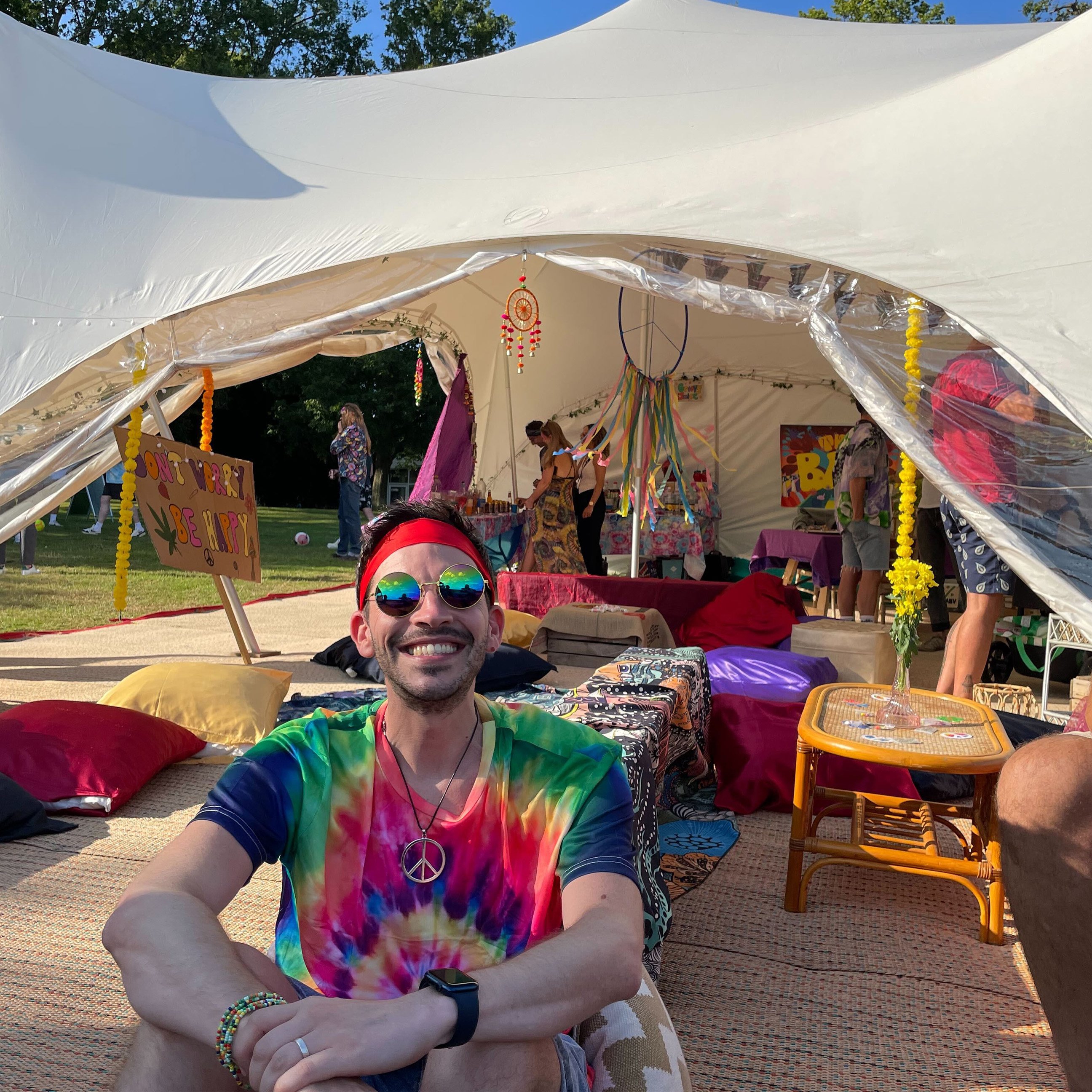 A person in a colourful tie-dye shirt and reflective sunglasses sitting on cushions in front of an outdoor festival-style tent decorated with signs, pillows, and hanging ornaments.