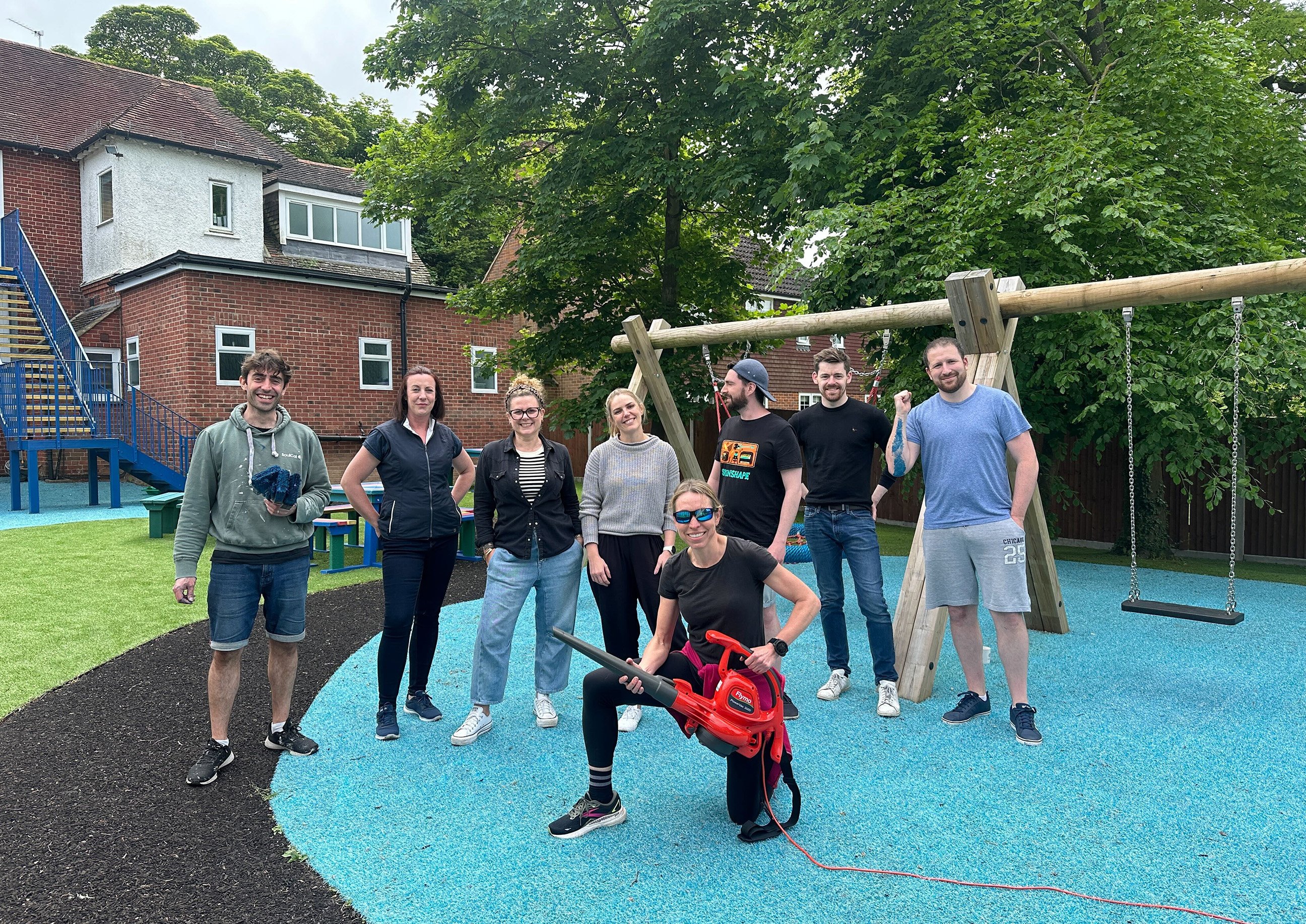 A group of volunteers posing in a playground they have been working on, with one person holding a leaf blower and others smiling behind them.