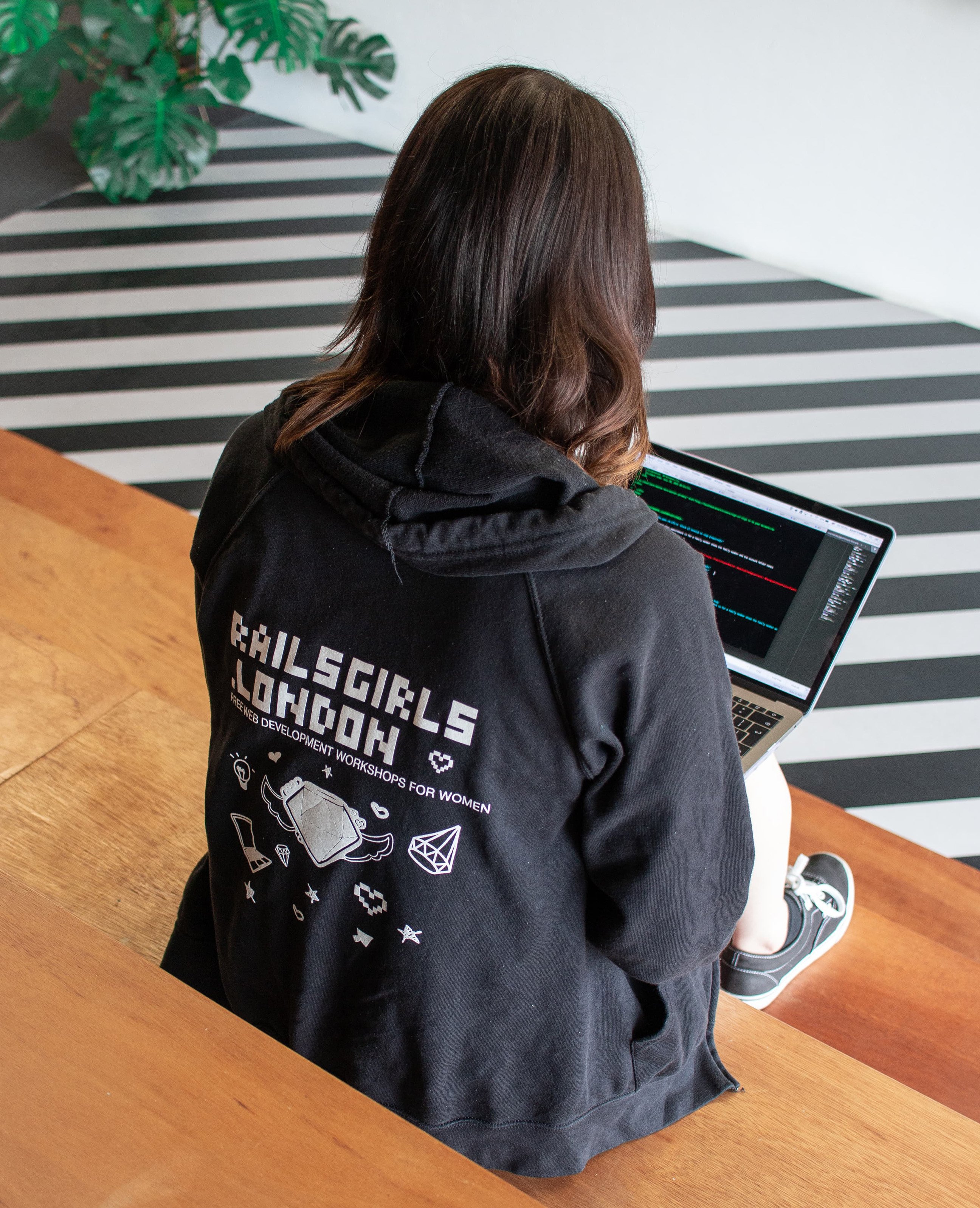 A woman sits on wooden steps with a laptop, wearing a black Rails Girls London hoodie, coding with a striped wall pattern in the background.