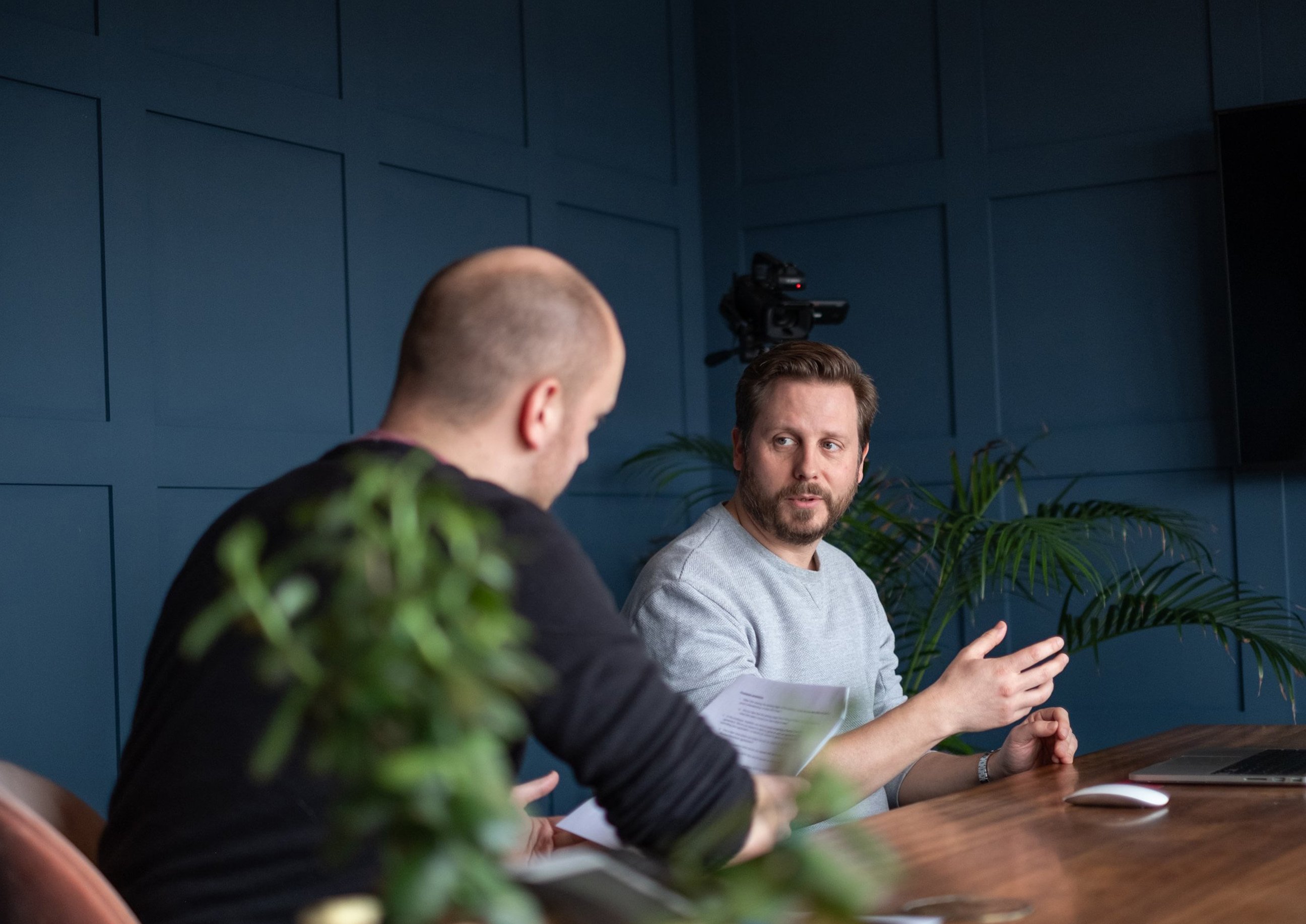 Two men sit at a table in a meeting room, discussing notes, with a camera and plants visible in the background.