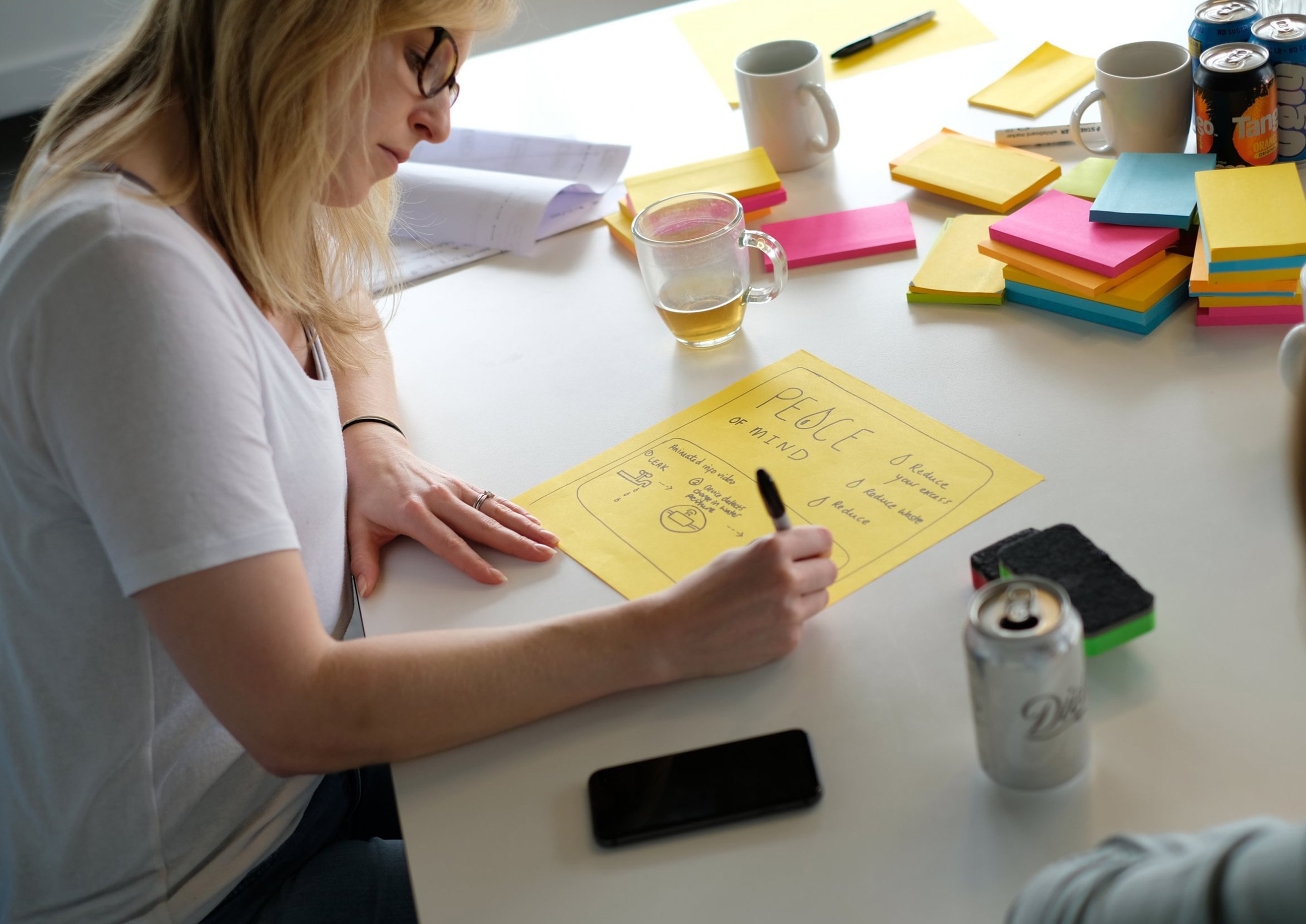 A woman writes on a large yellow sheet during a workshop, surrounded by colourful sticky notes, mugs, and drinks on the table.