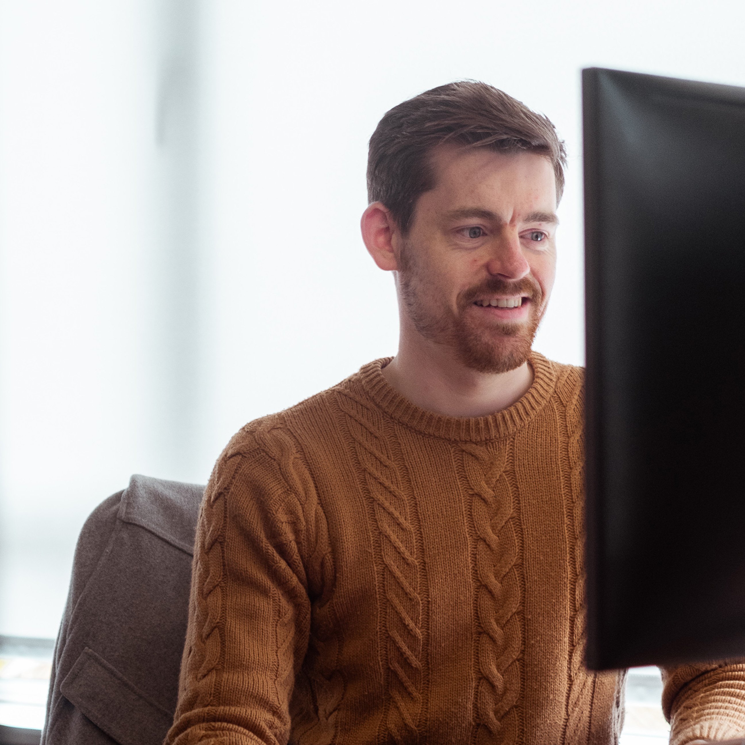 A man wearing a brown cable-knit jumper works at his computer, smiling as he looks at the screen.