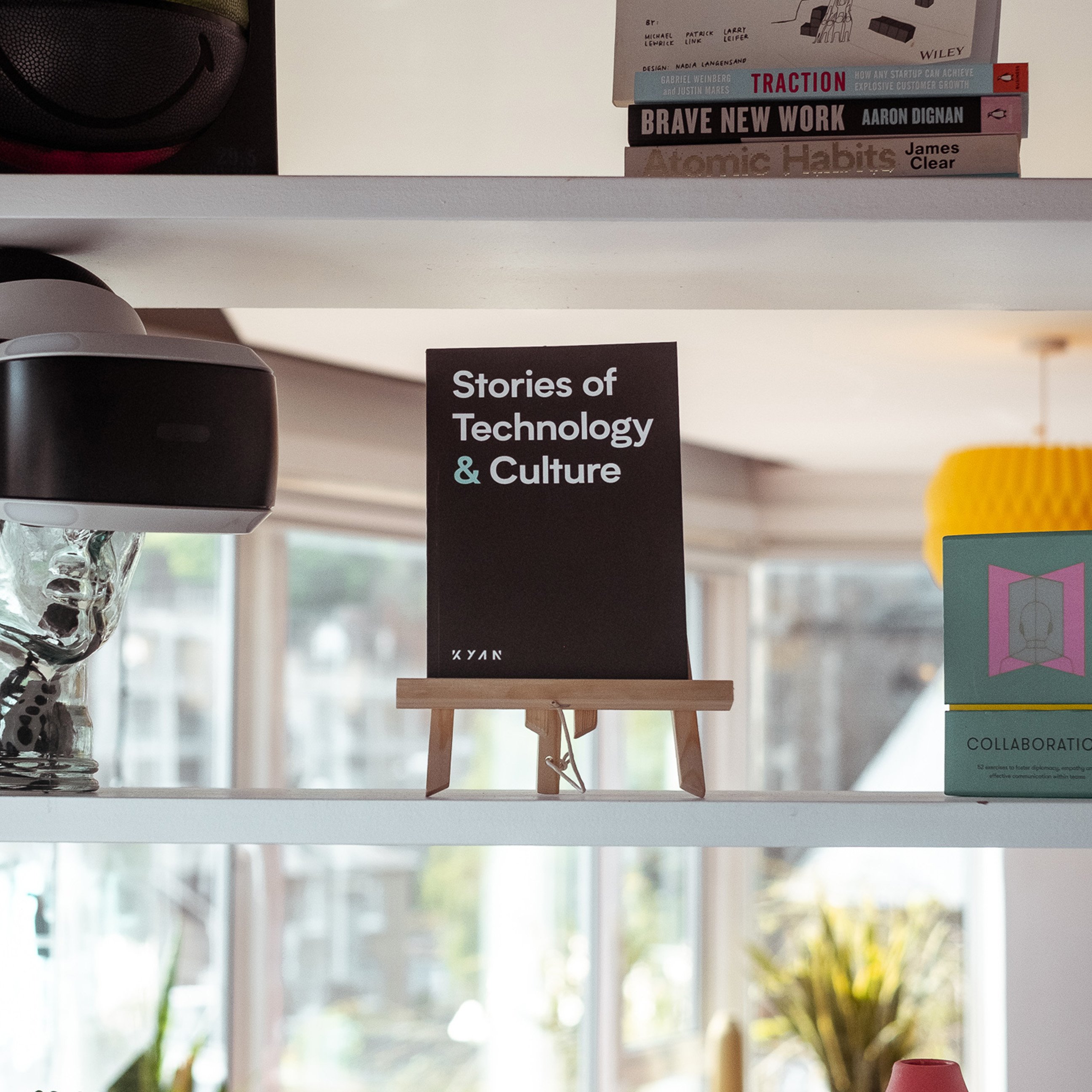A small easel displaying a book titled “Stories of Technology & Culture” on a bright office shelf, surrounded by books, decor items, and a VR headset.