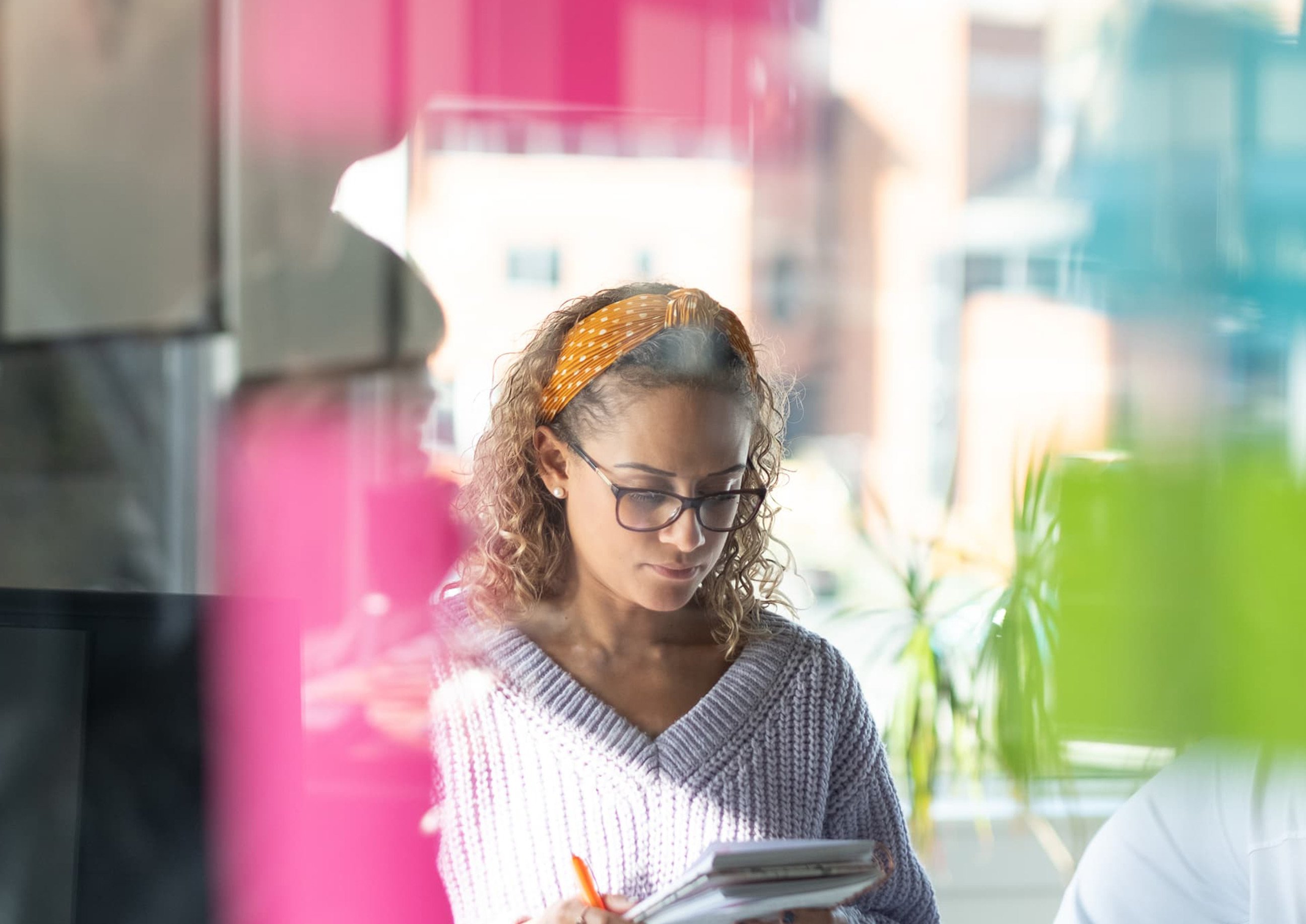 A woman wearing glasses and an orange polka-dot headband writes in a notebook, seen through a window covered with colorful sticky notes in a bright office space.