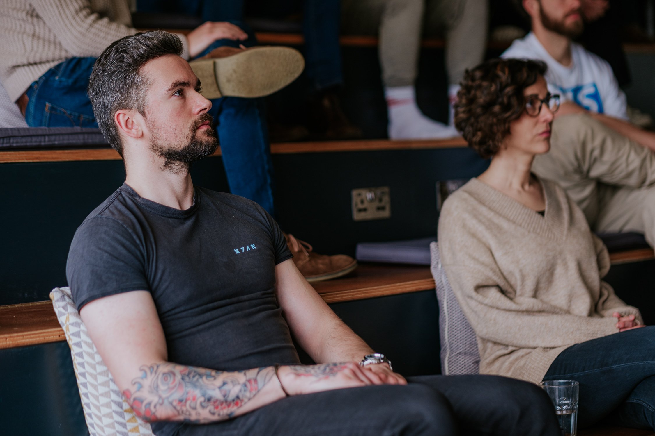 A group of people sit on tiered seating during a talk, with a man in a dark T-shirt and a woman in a beige sweater focused on the presentation.