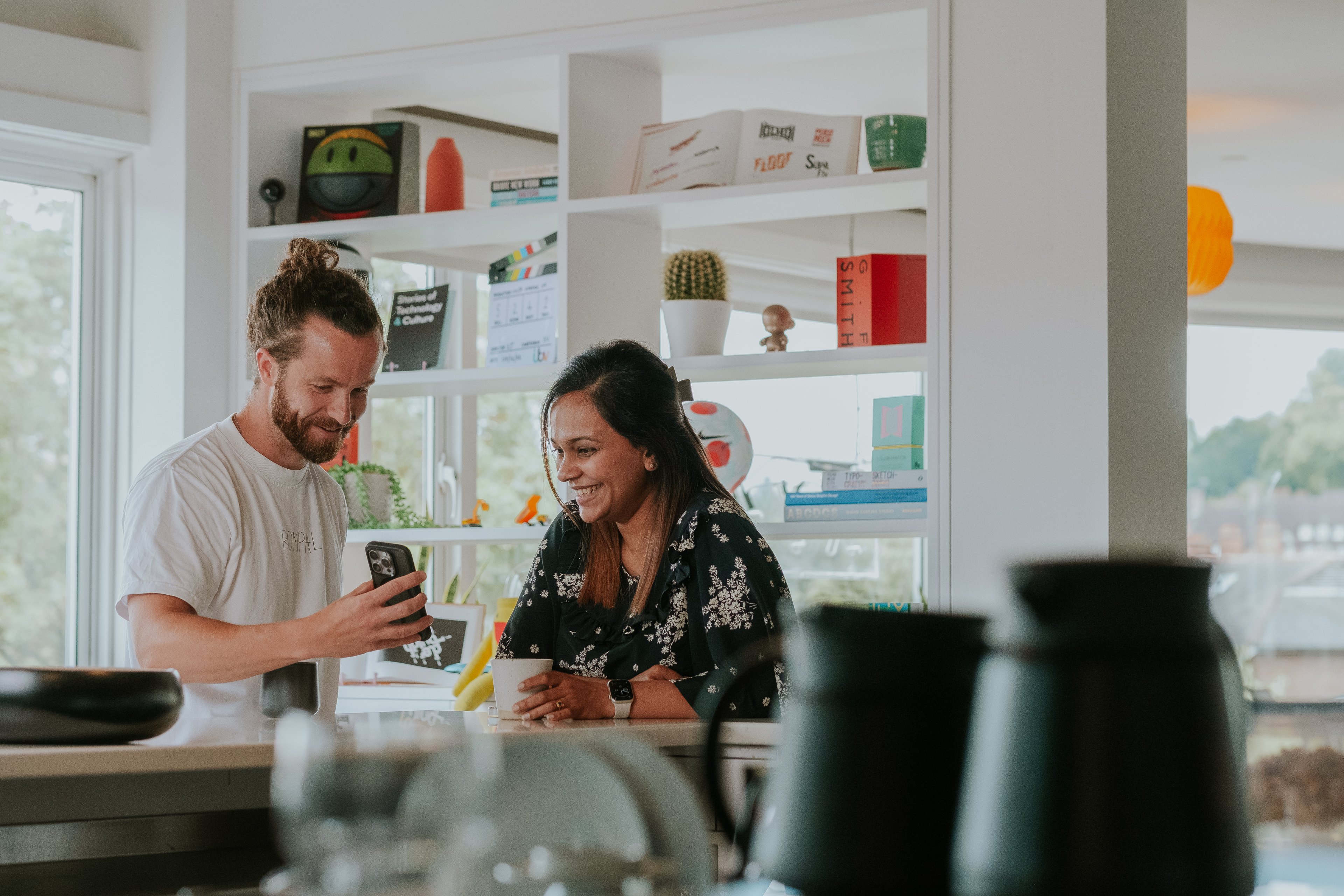 A man and woman stand in a bright kitchen area smiling and looking at something on a smartphone together.