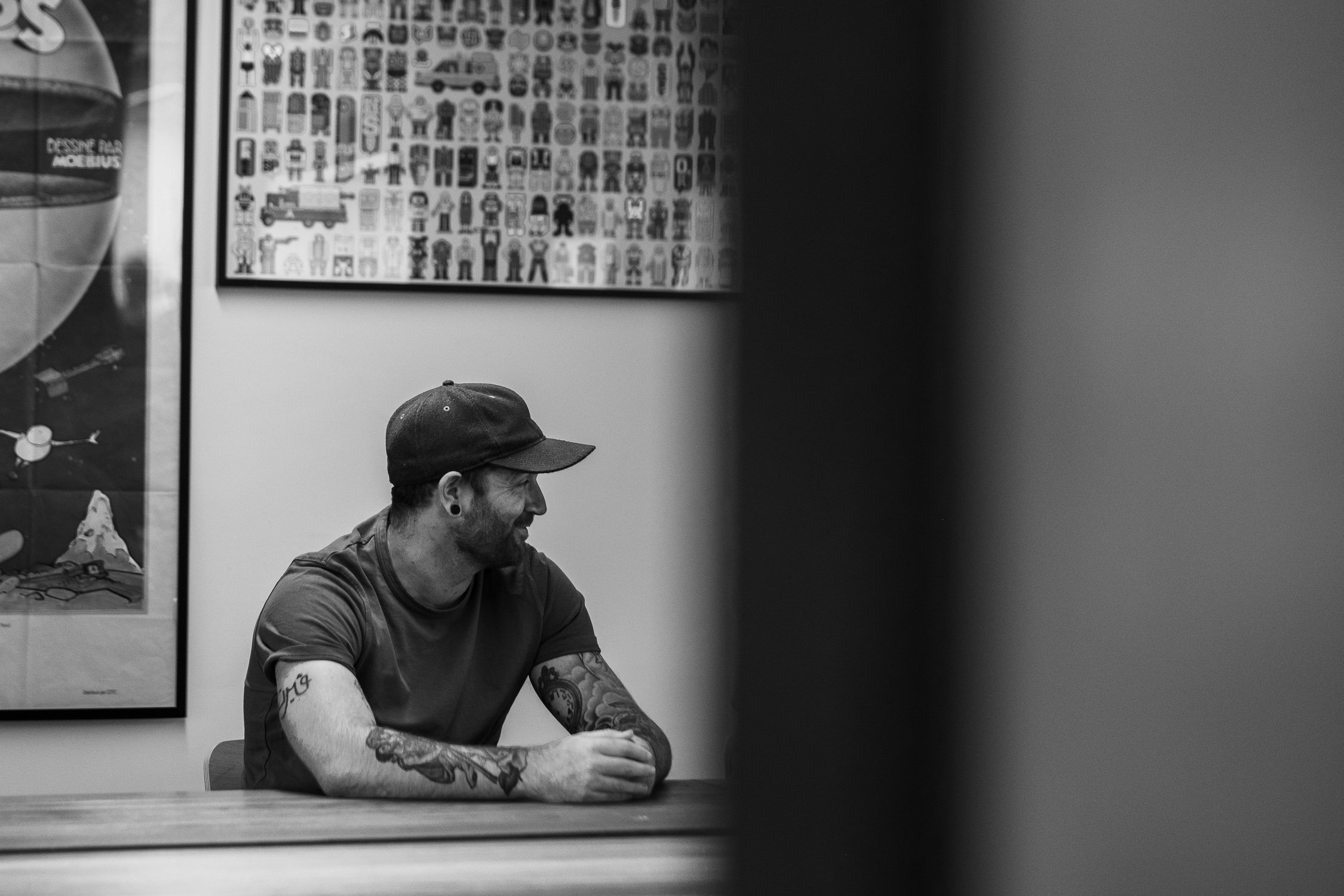 A man with tattoos and a cap sits at a table in a room decorated with framed posters, smiling as he looks to the side.