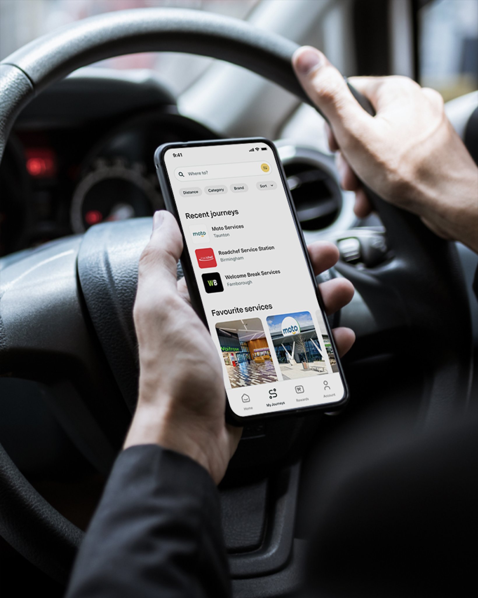 Driver holding a smartphone while sitting behind the wheel, viewing the Pitstop app's home screen showing recent journeys and service station options.