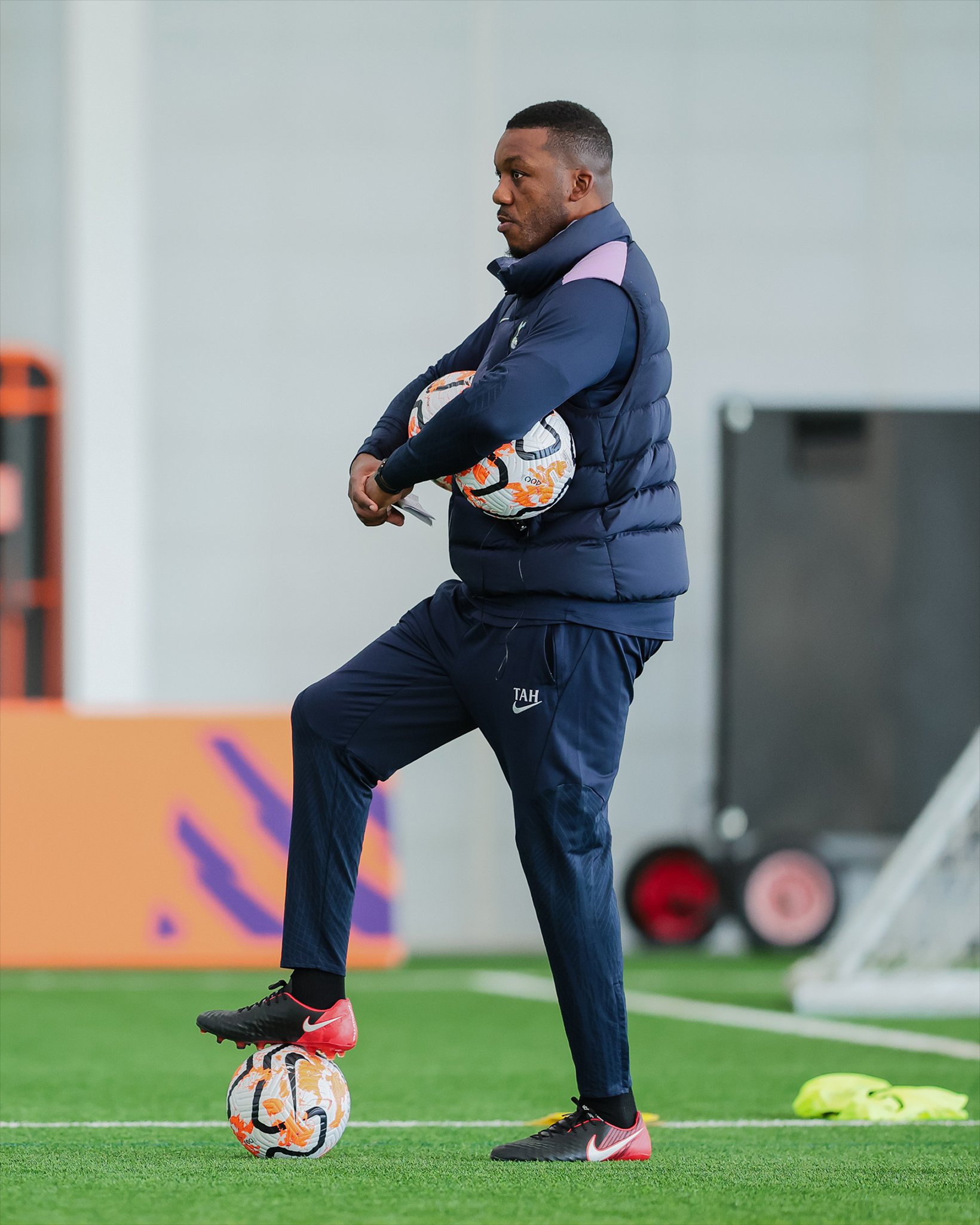 A football coach in training gear balances one foot on a football while holding two more balls under his arm on an indoor pitch.