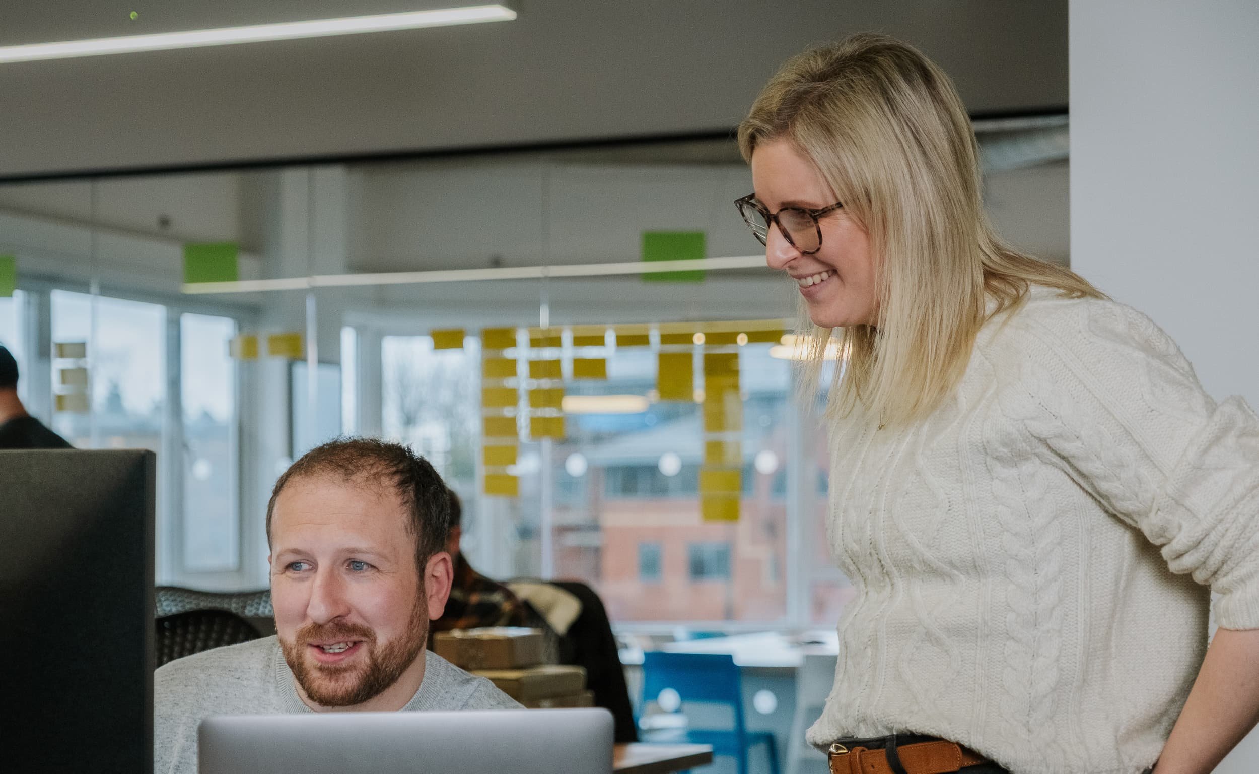A woman stands beside a seated colleague, smiling as they review content together on a desktop computer in a bright office.