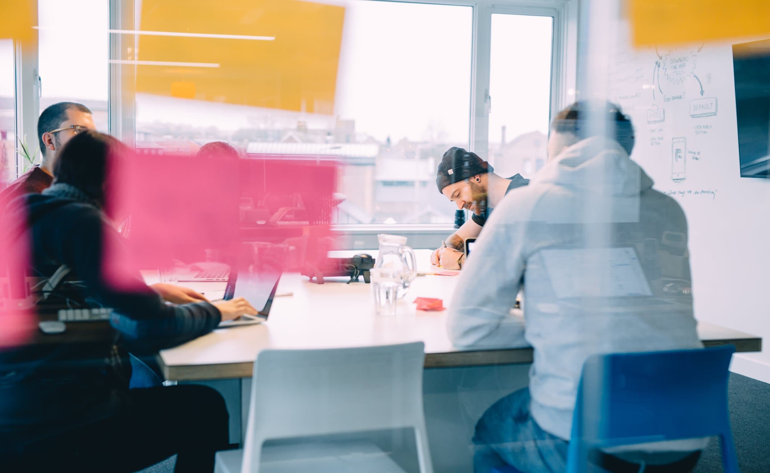 A team collaborates in a meeting room, partially obscured by brightly coloured sticky notes stuck to the glass walls. One person writes on a laptop while another sketches.