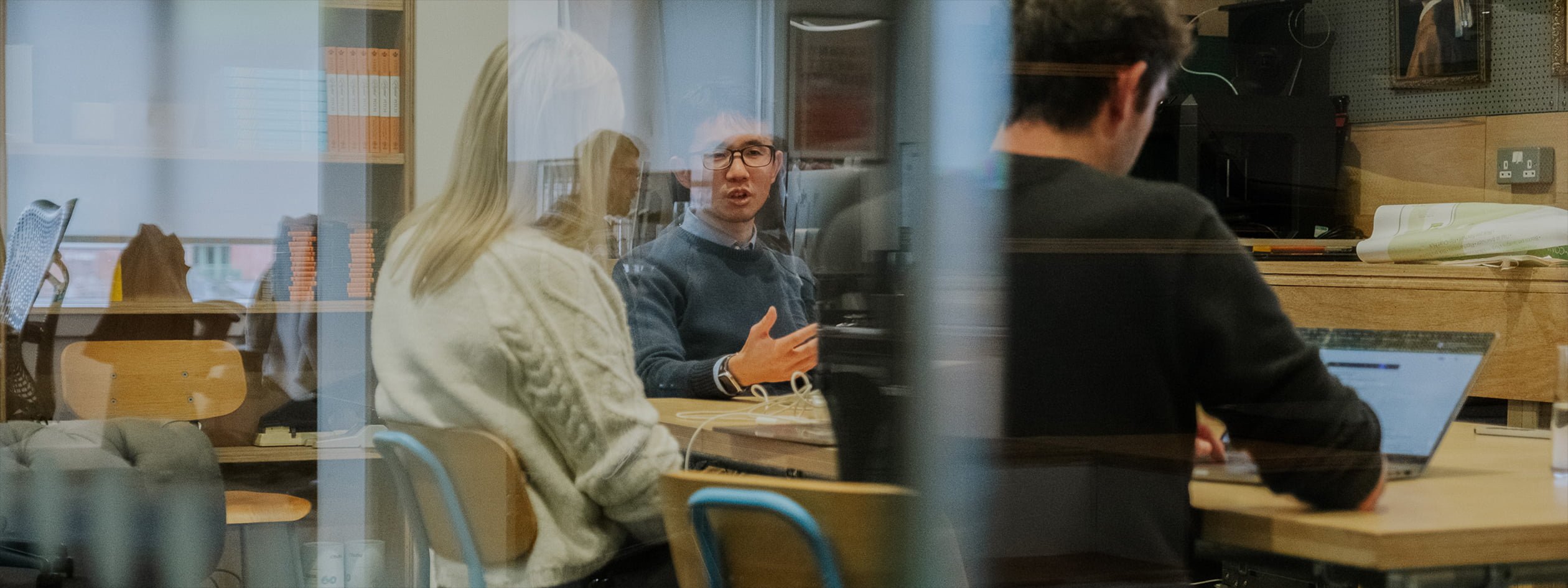 People sit around a meeting table in a modern office, collaborating with laptops. The scene is photographed through glass with reflections of the external office environment visible.