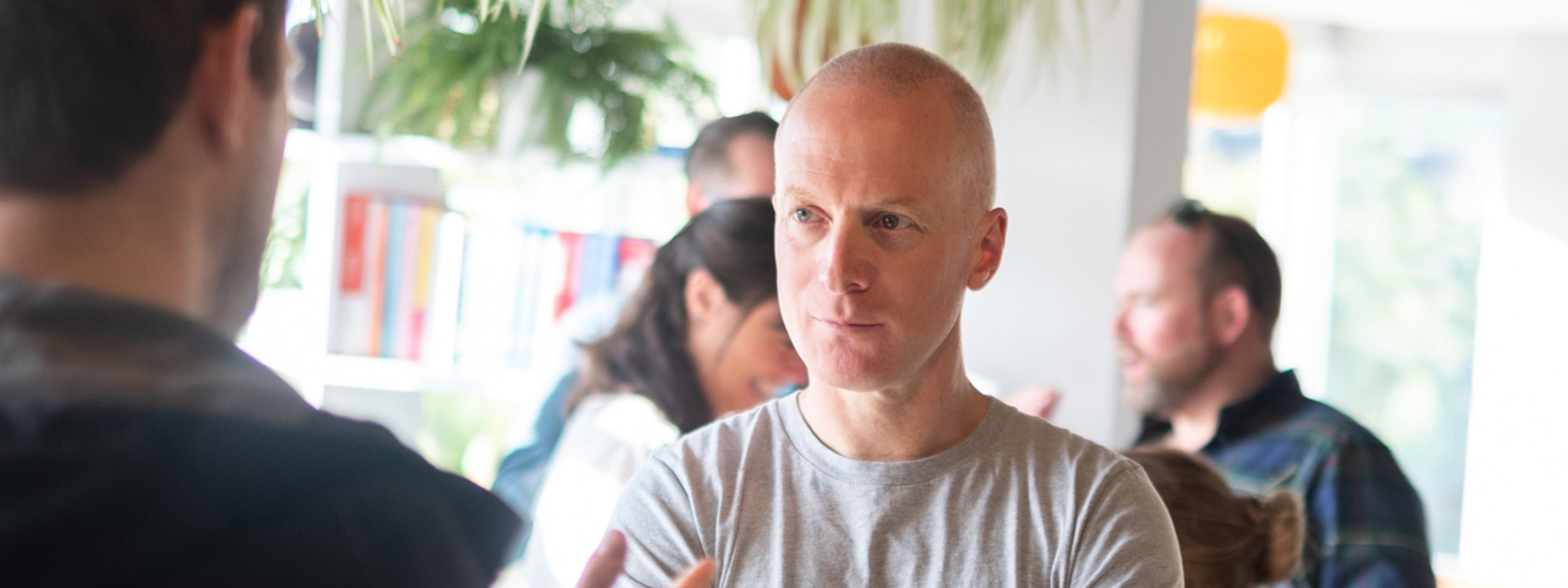 A man in a grey t-shirt listens attentively during a conversation in a bright, plant-filled office, with colleagues talking in the background.